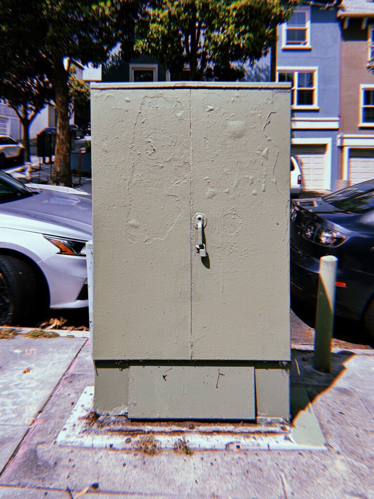 A light gray utility cabinet mounted on a concrete base along a San Francisco sidewalk, enclosing electrical or communications infrastructure behind locked metal doors.