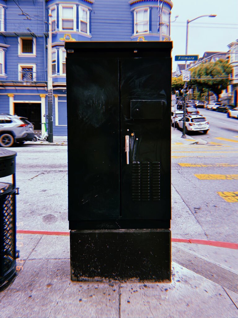 A black utility cabinet installed on a San Francisco street corner, enclosing electrical or communications infrastructure behind secure metal access doors.