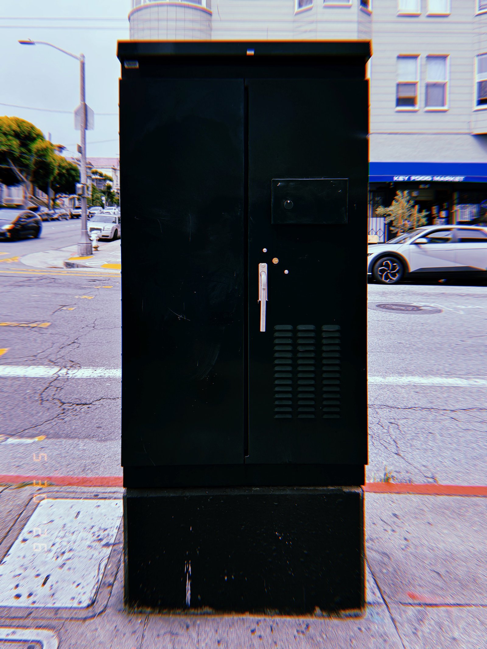 A black utility cabinet positioned at a San Francisco street intersection, housing electrical or communications infrastructure behind secure metal access doors.