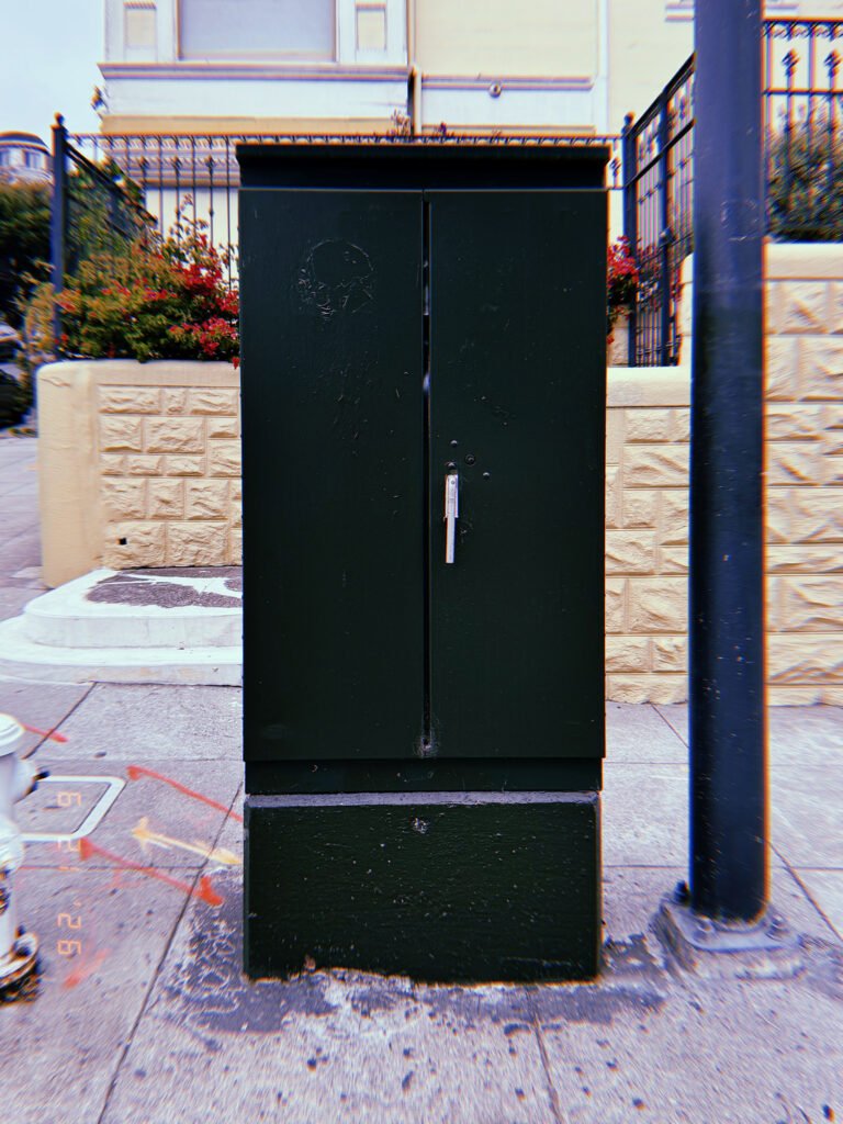 A dark green utility cabinet mounted on a concrete base beside a sidewalk. The metal enclosure protects electrical or communications infrastructure and provides secure maintenance access.