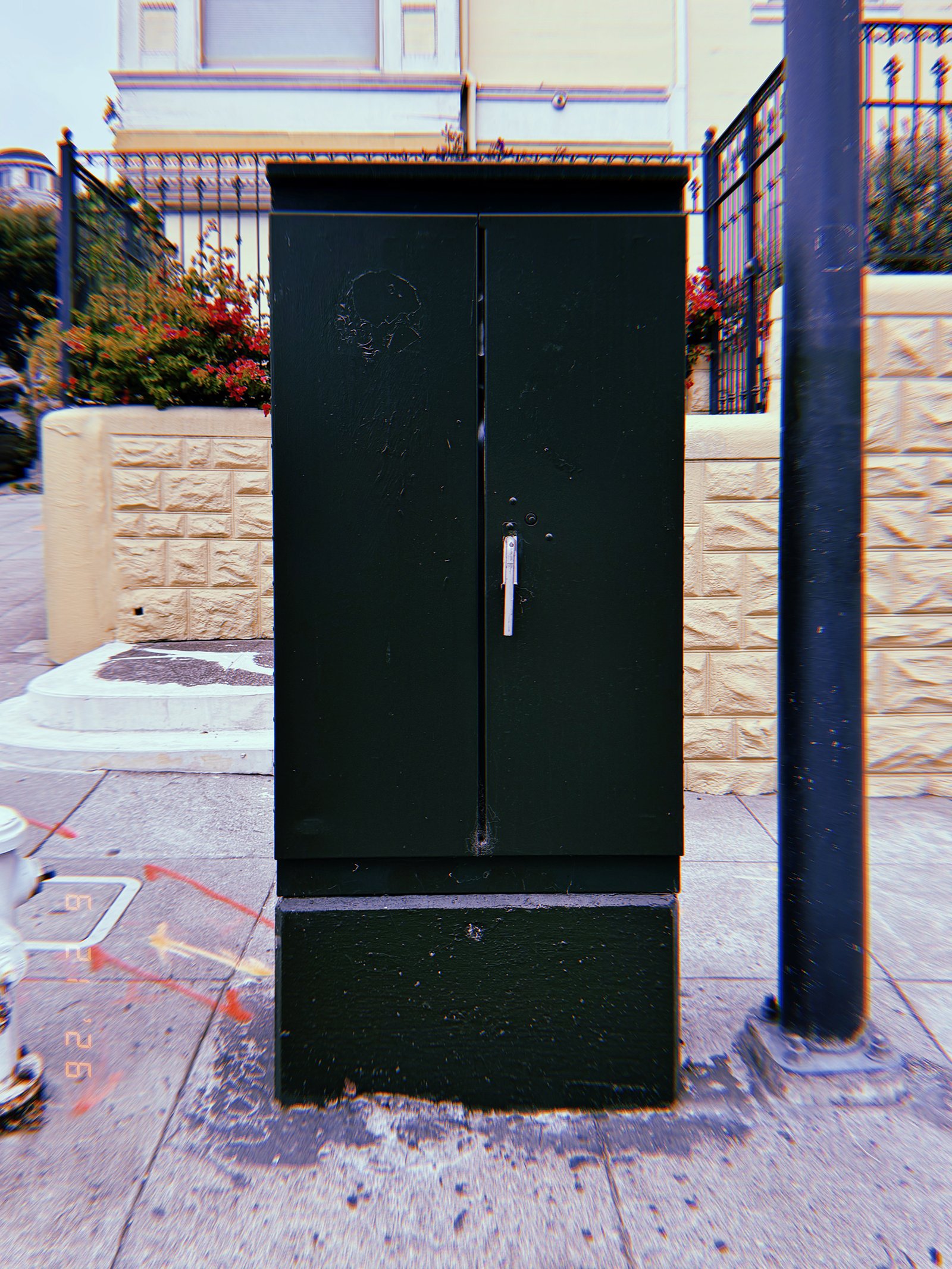 A dark green utility cabinet mounted on a concrete base beside a sidewalk. The metal enclosure protects electrical or communications infrastructure and provides secure maintenance access.