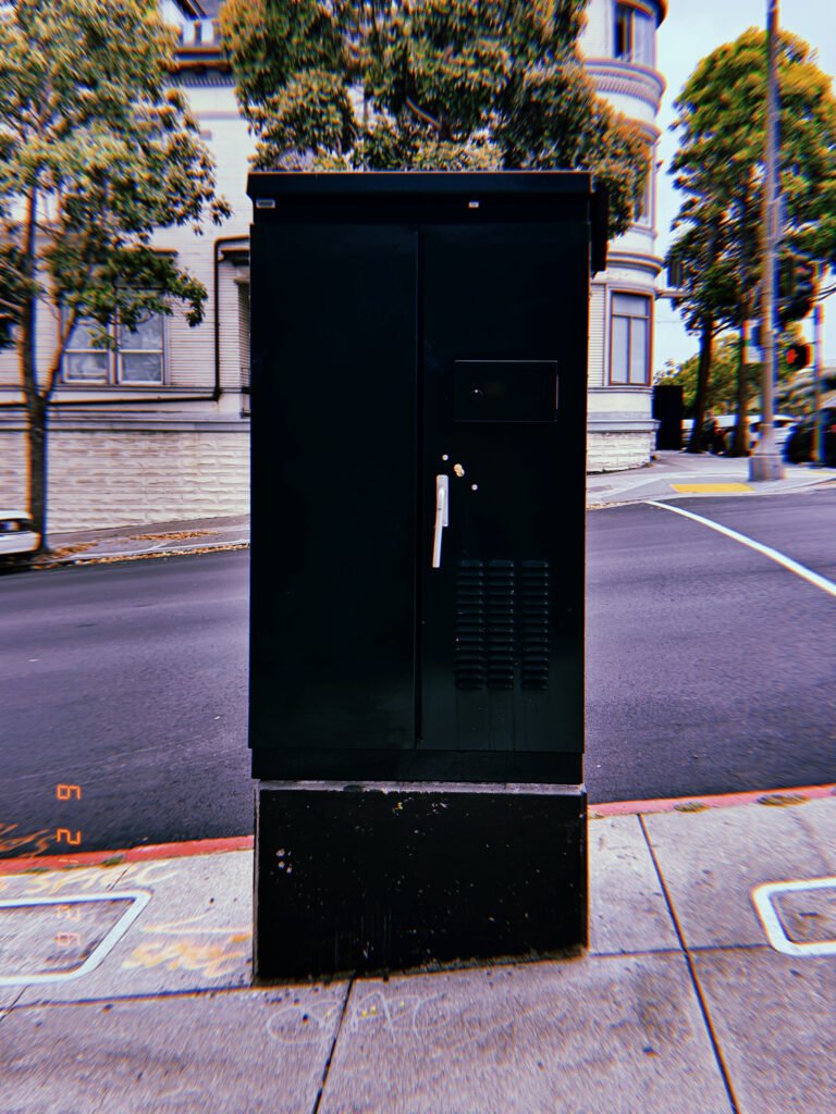 A black utility cabinet installed on a concrete base at a street corner. The freestanding enclosure provides protected access to electrical or communications infrastructure within the urban network.