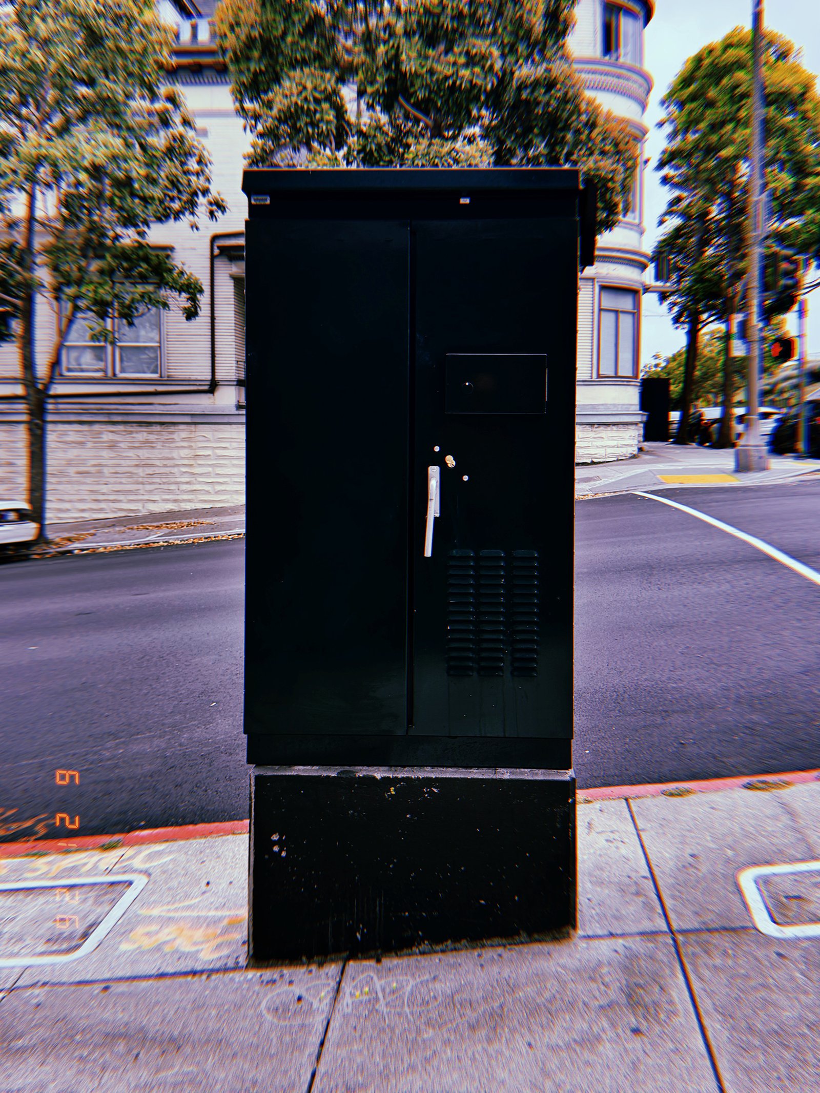 A black utility cabinet installed on a concrete base at a street corner. The freestanding enclosure provides protected access to electrical or communications infrastructure within the urban network.