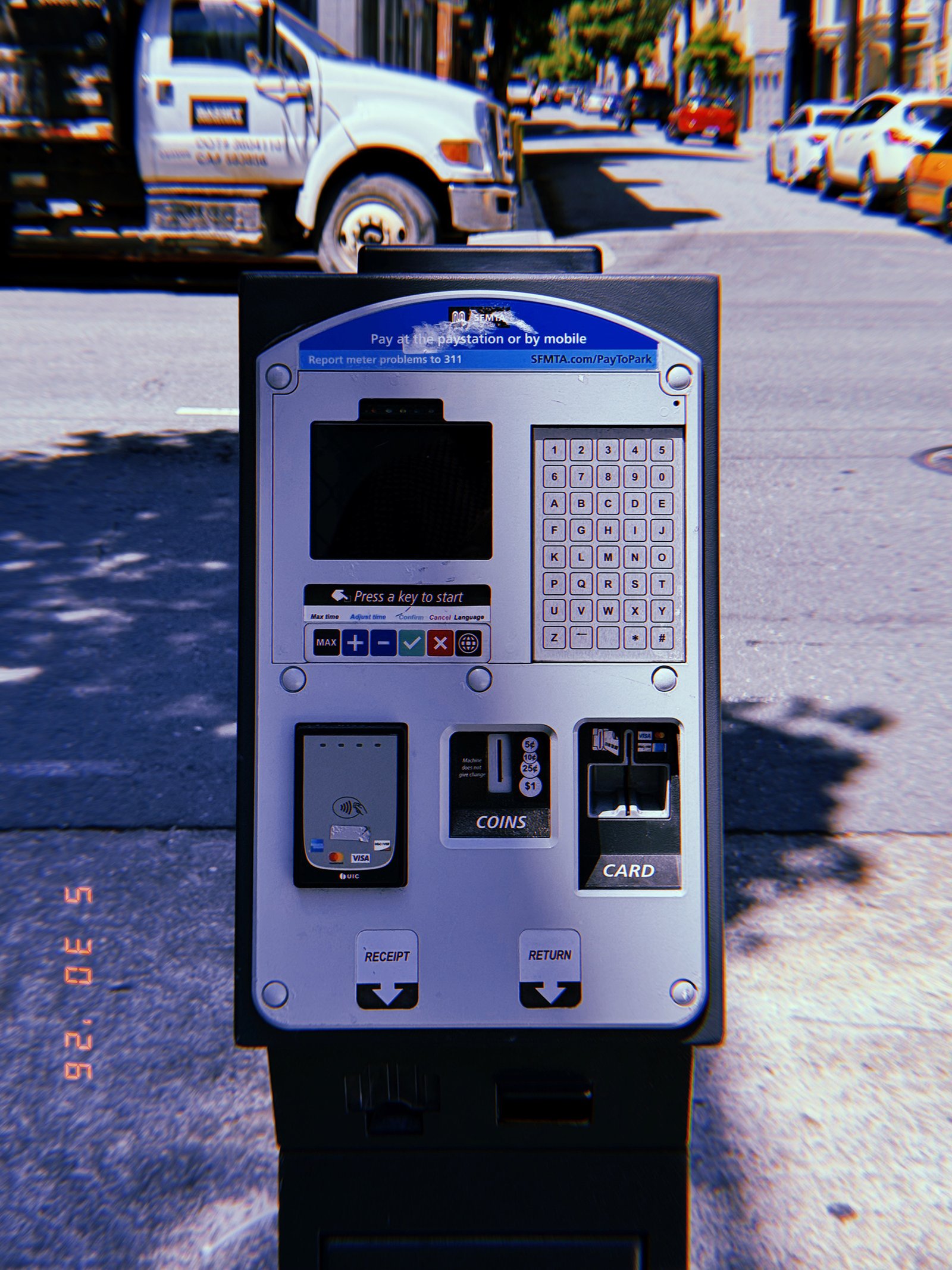 A San Francisco parking payment kiosk photographed frontally at the edge of a residential street, showing its digital display, keypad, contactless payment reader, coin slot, and card payment terminal.