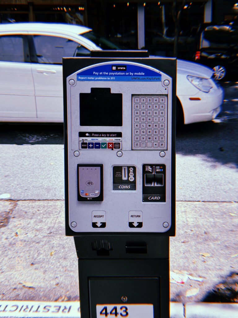 A San Francisco parking payment kiosk with touchscreen, keypad, contactless payment reader, coin slot, and card terminal standing beside the curb with parked vehicles in the background.