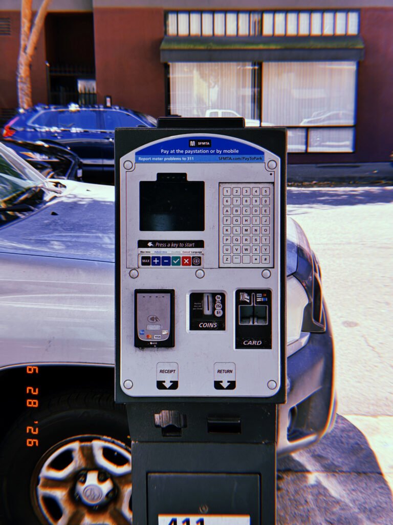 A San Francisco parking payment kiosk with digital display, keypad, contactless payment reader, coin slot, and card terminal positioned in front of parked vehicles.