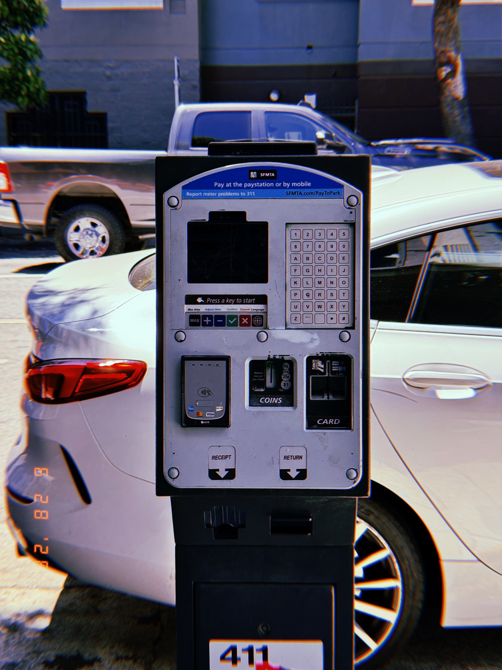A San Francisco parking payment kiosk with digital display, keypad, contactless payment reader, coin slot, and card terminal standing in front of parked vehicles.