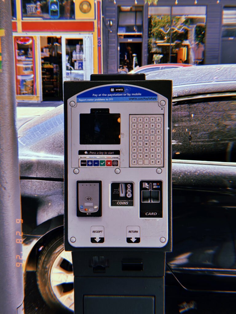 A San Francisco parking payment kiosk with touchscreen, keypad, contactless reader, coin slot, and card terminal installed along a commercial street with parked vehicles.