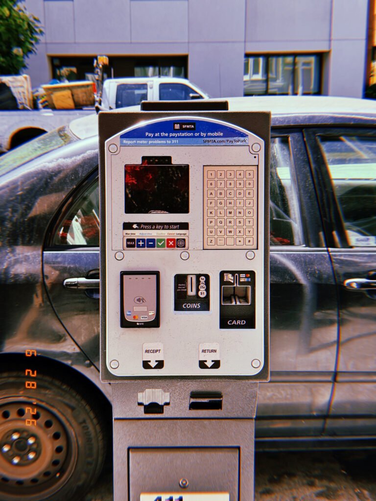 A San Francisco parking payment kiosk with digital display, keypad, contactless payment reader, coin slot, and card terminal positioned in front of a parked vehicle.