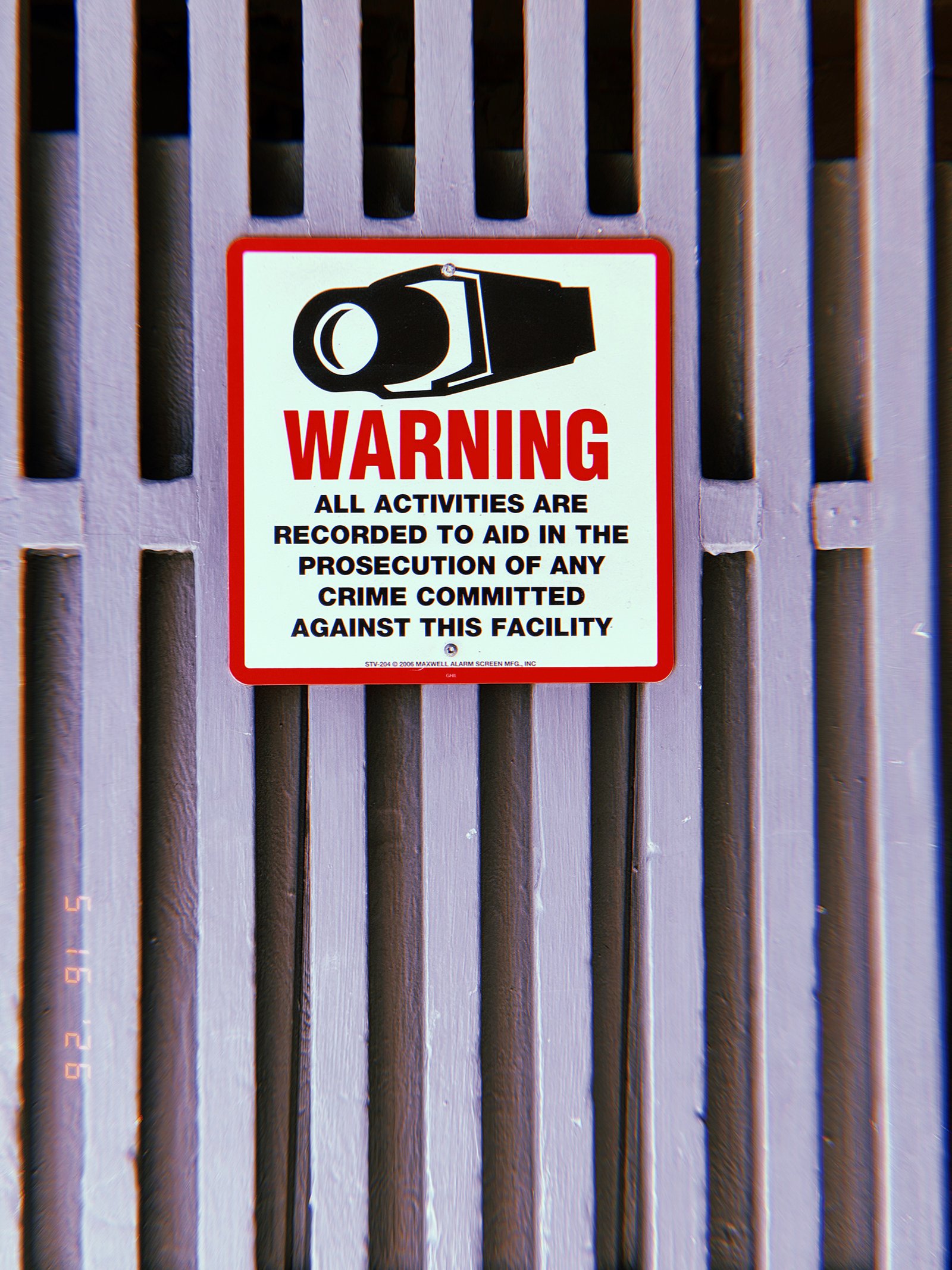 Warning sign with a security camera graphic stating that all activities are recorded, mounted on a gray metal gate in San Francisco.