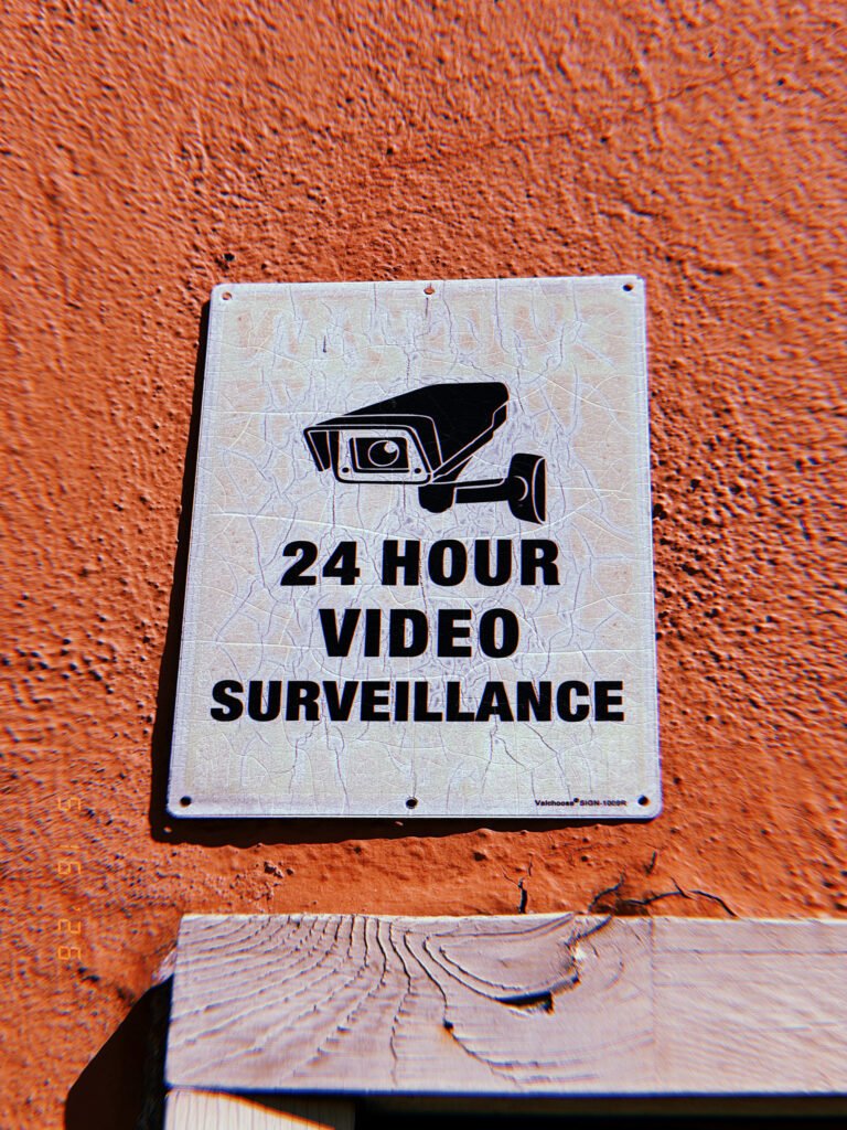 Weathered 24-hour video surveillance sign with a security camera graphic mounted on an orange exterior wall in San Francisco.