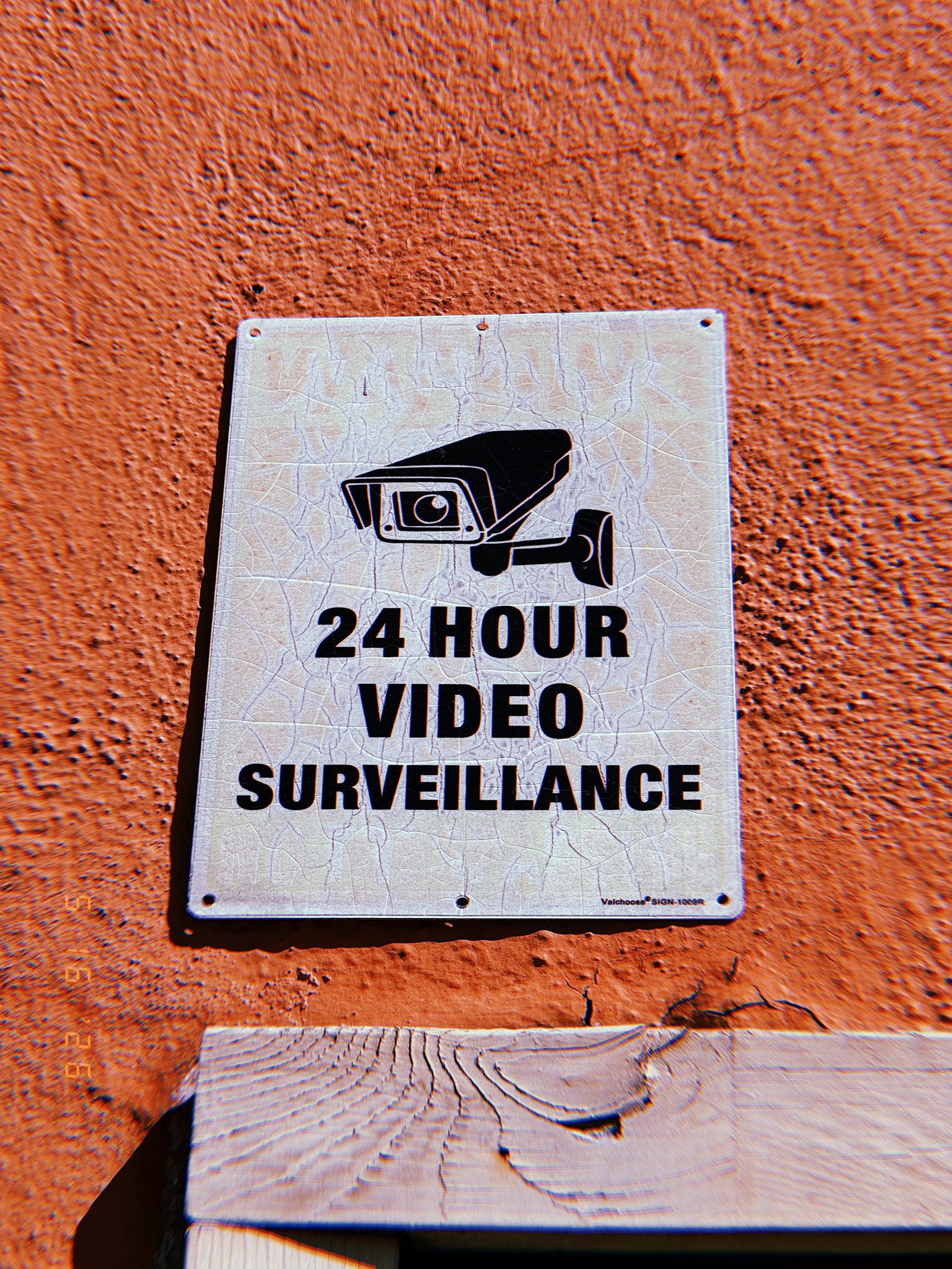 Weathered 24-hour video surveillance sign with a security camera graphic mounted on an orange exterior wall in San Francisco.