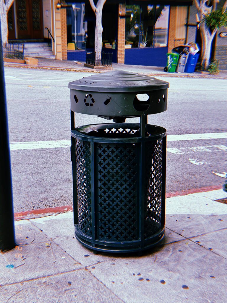 Black public recycling receptacle on a San Francisco sidewalk, with residential recycling bins and storefronts across the street.