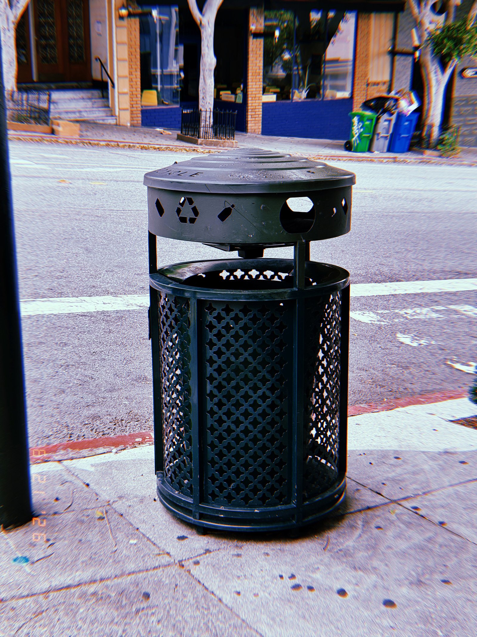 Black public recycling receptacle on a San Francisco sidewalk, with residential recycling bins and storefronts across the street.