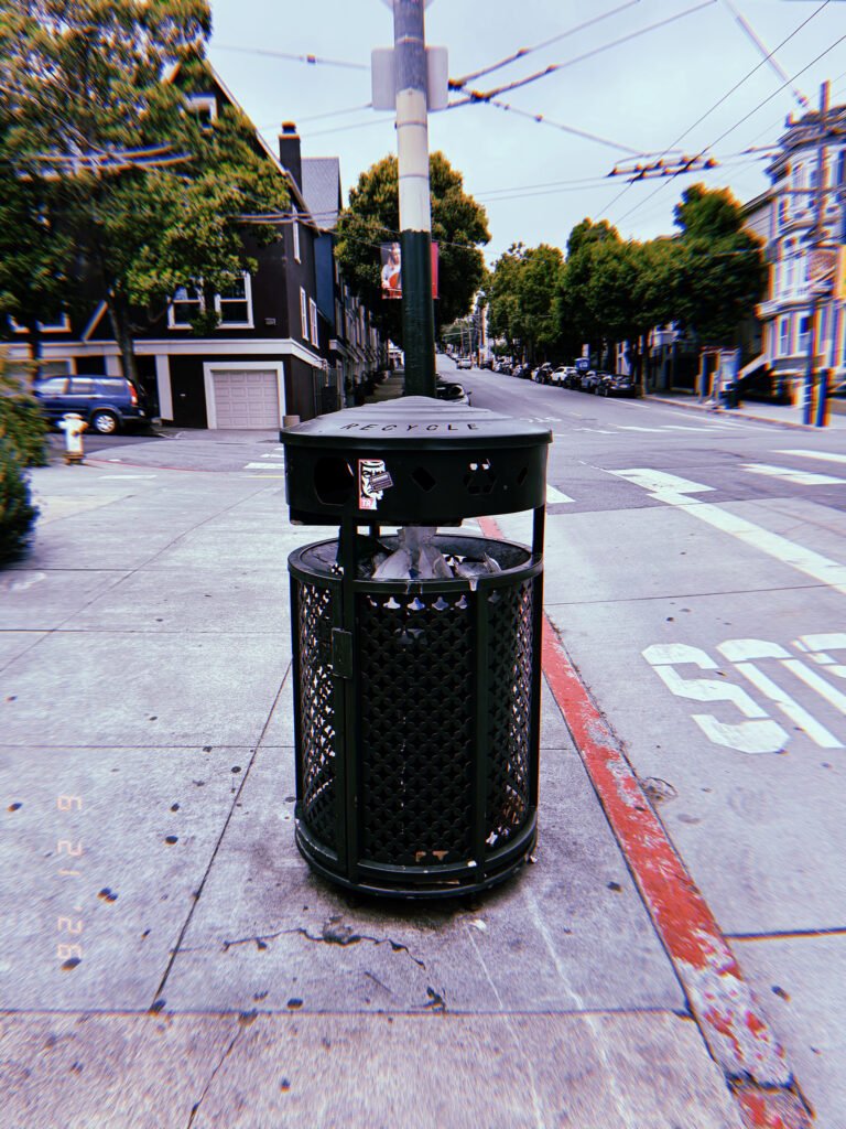 Black public recycling receptacle on a San Francisco sidewalk at an intersection, with residential buildings and overhead transit wires behind it.