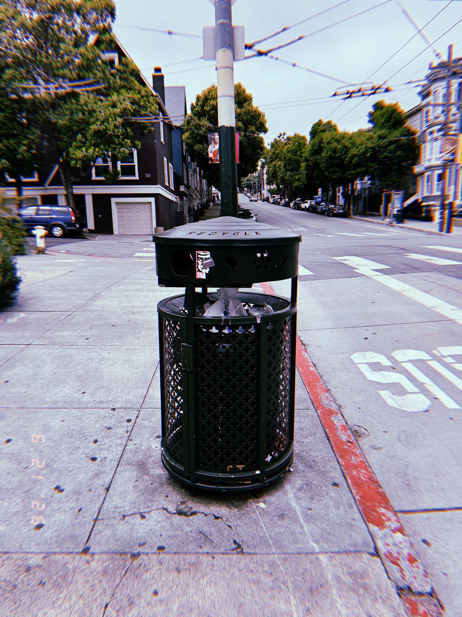 Black public recycling receptacle on a San Francisco sidewalk at an intersection, with residential buildings and overhead transit wires behind it.