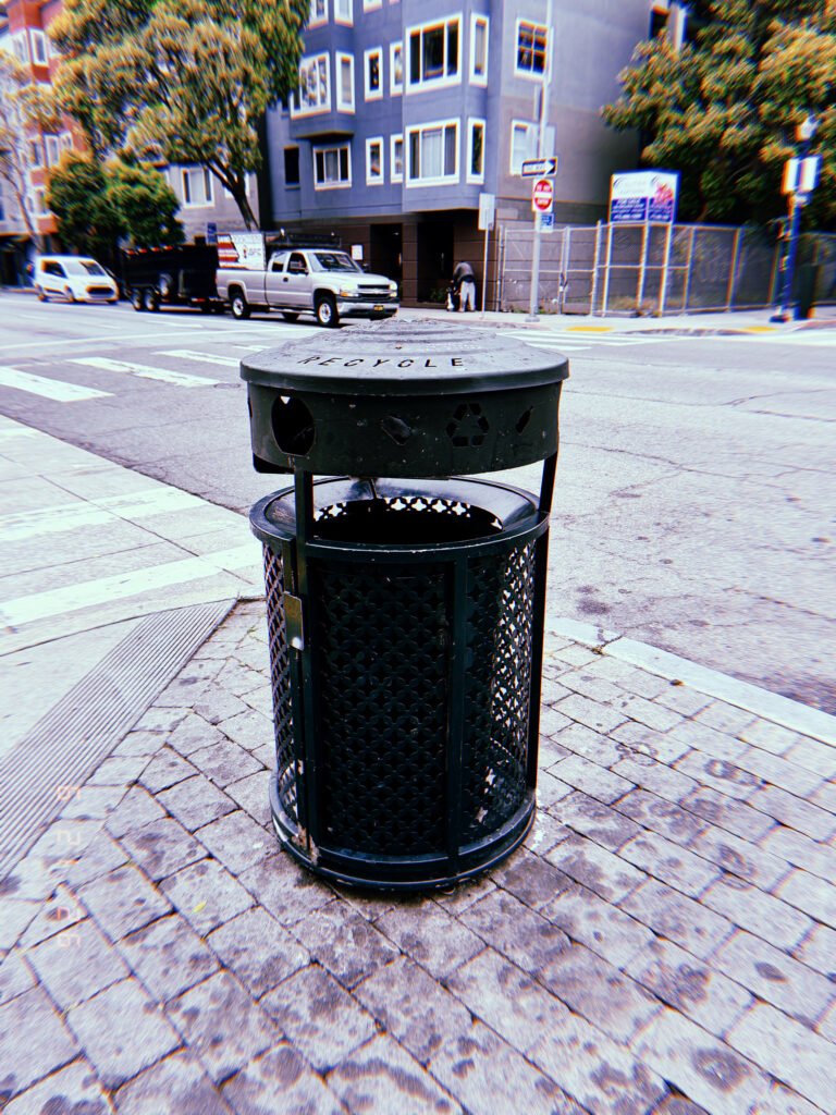 Black public recycling receptacle on a paved corner at a San Francisco intersection, with residential buildings and vehicles behind it.