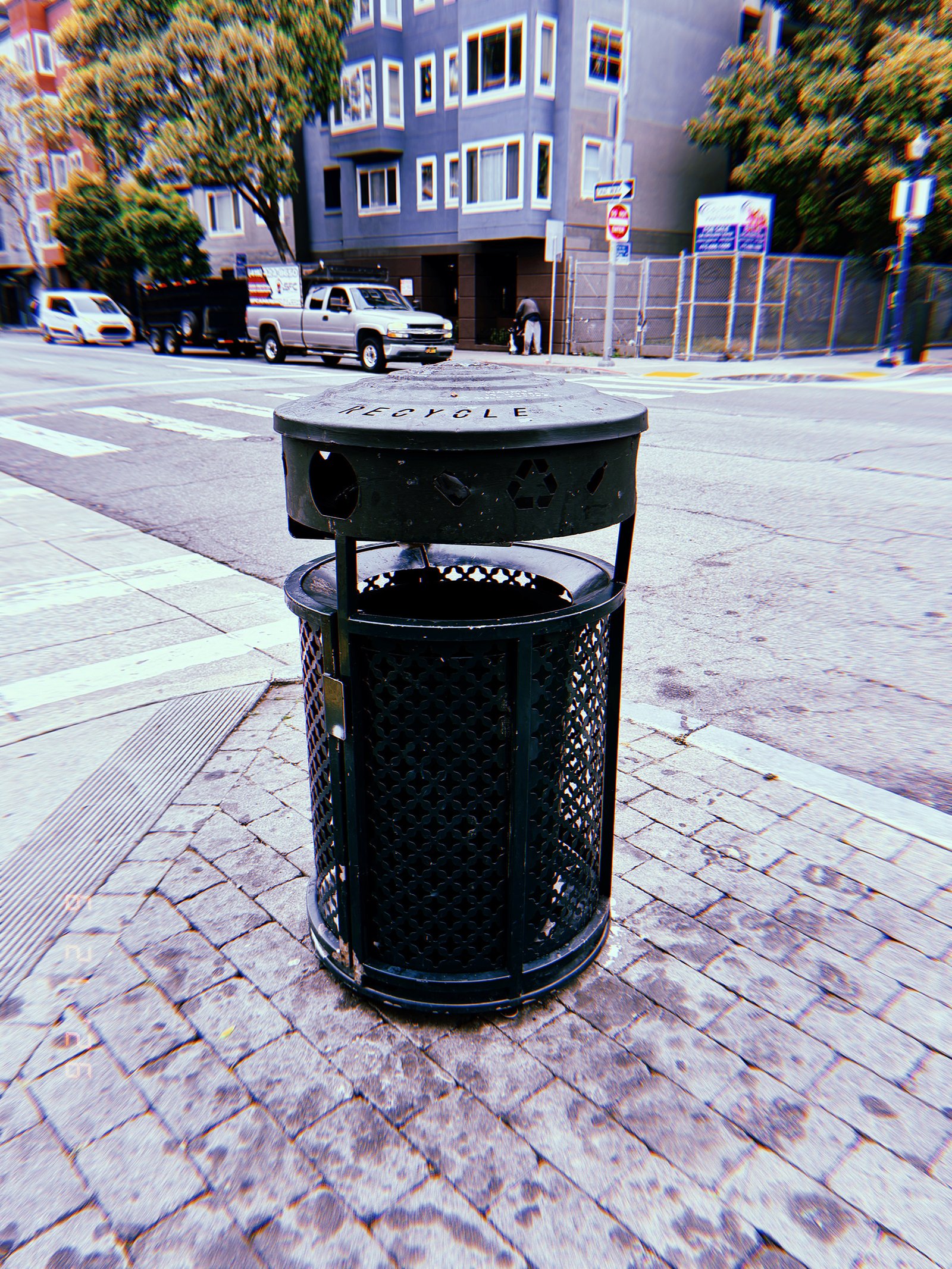 Black public recycling receptacle on a paved corner at a San Francisco intersection, with residential buildings and vehicles behind it.