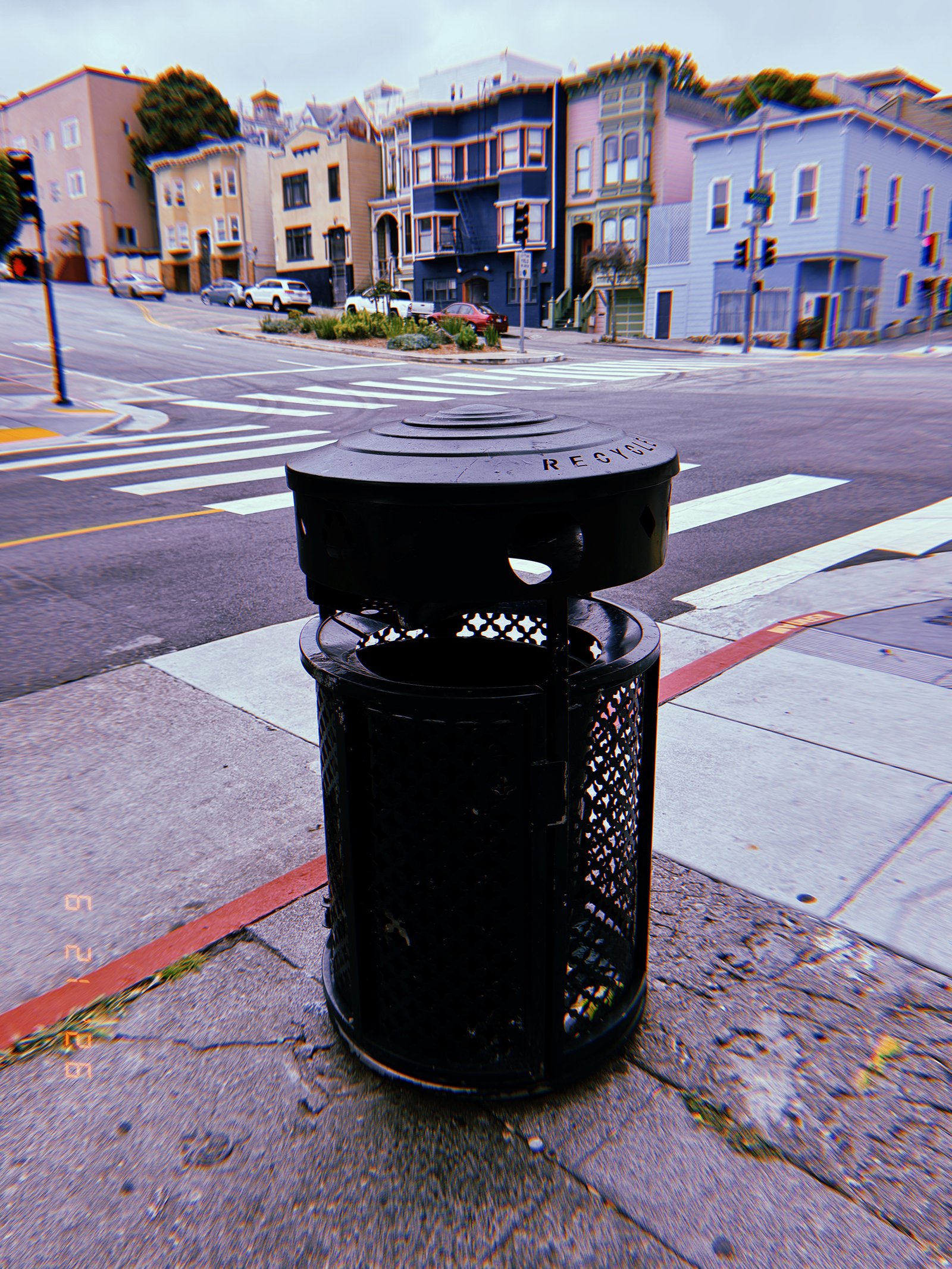 Black public recycling receptacle on a San Francisco street corner, with crosswalks and residential buildings in the background.