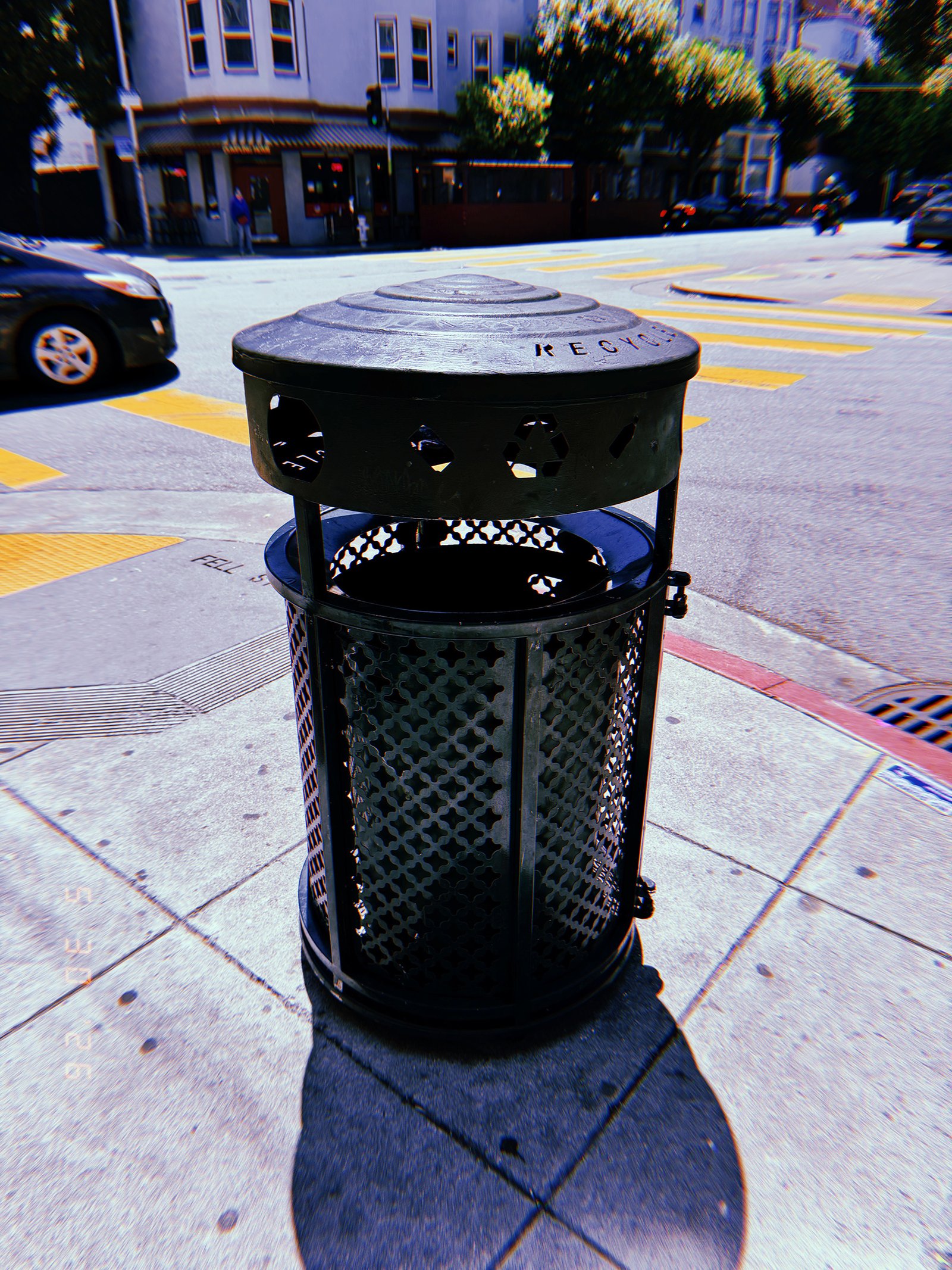 Black public recycling receptacle on a San Francisco street corner, with yellow crosswalk markings and buildings in the background.