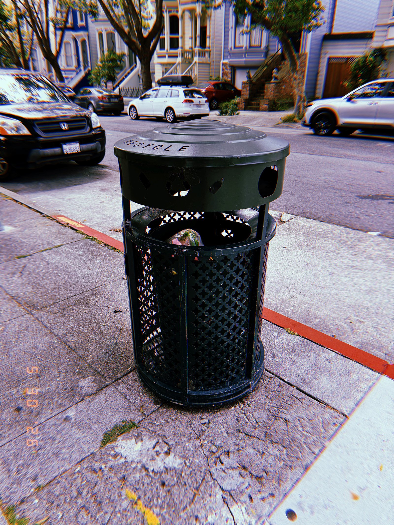 Black public recycling receptacle on a San Francisco sidewalk, with parked cars and residential buildings in the background.