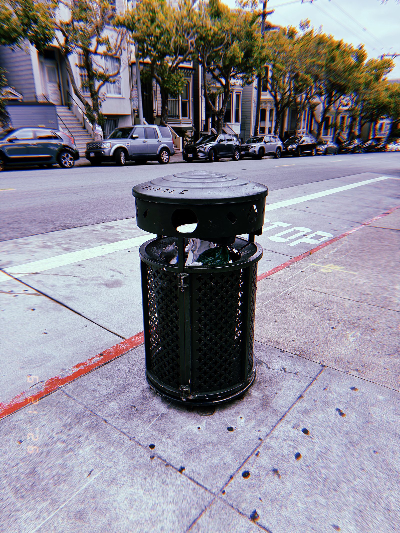 Black public recycling receptacle with visible waste inside, positioned on a sidewalk beside a residential street in San Francisco.