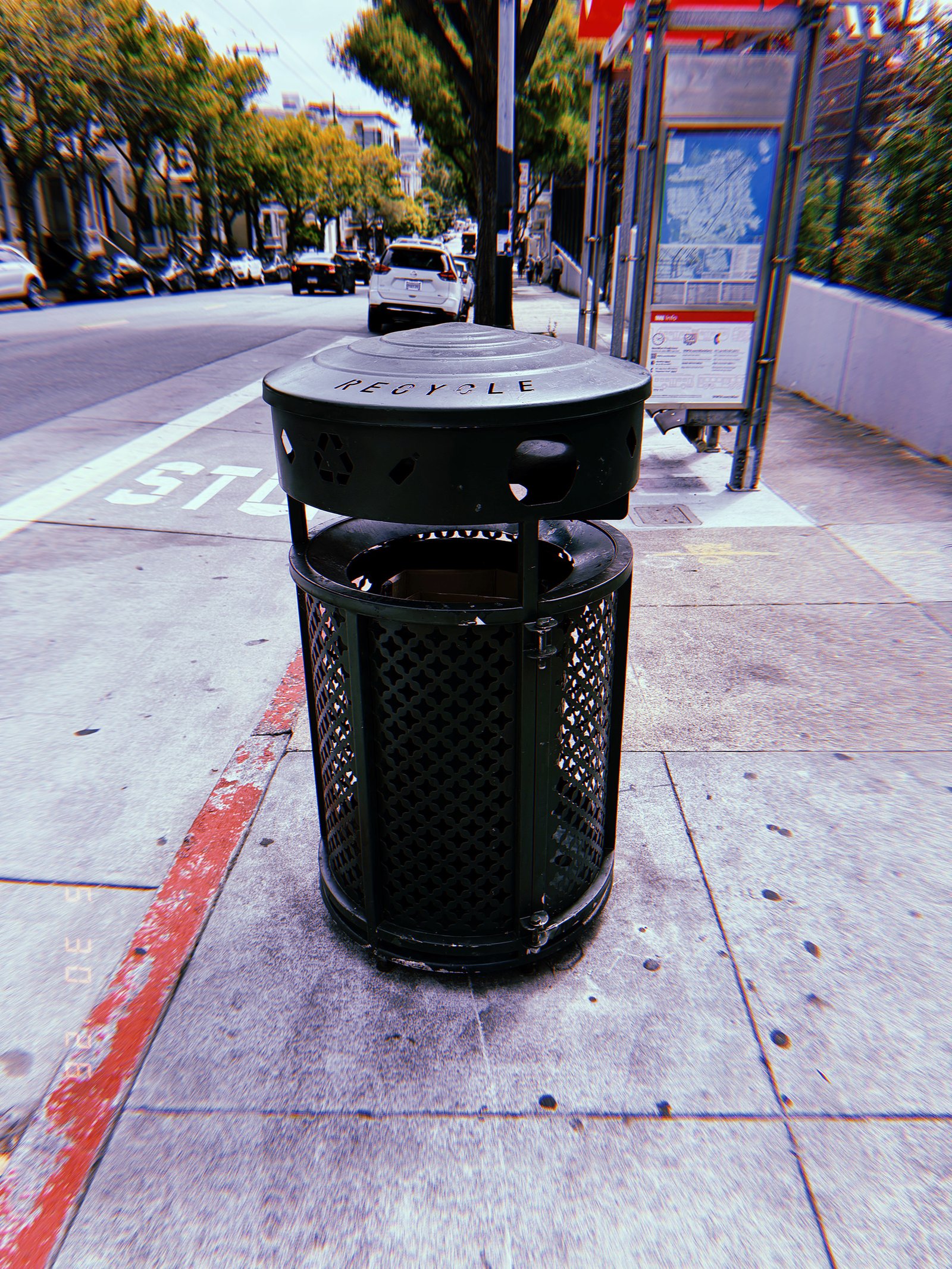 Black public recycling receptacle with a covered lid on a sidewalk beside a bus stop and city street in San Francisco.