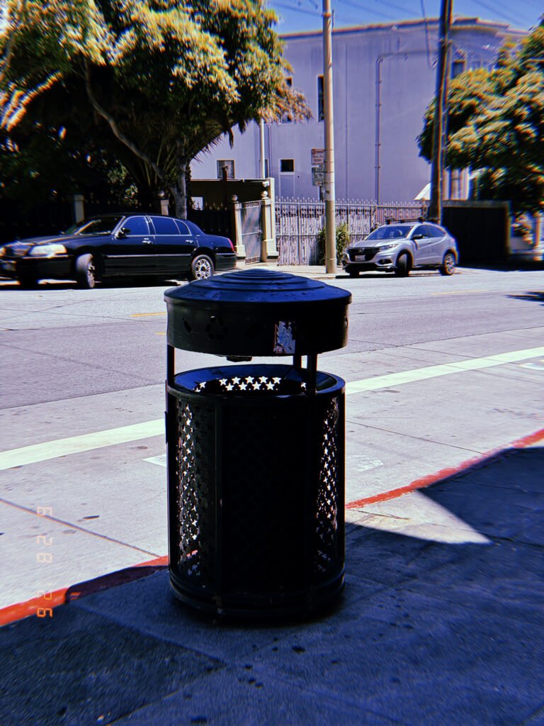 Black public trash can with a covered lid on a sidewalk beside a city street in San Francisco.