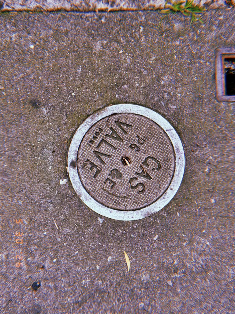 Round metal gas valve cover marked “VALVE GAS” embedded in a textured San Francisco sidewalk and surrounded by a pale circular border.