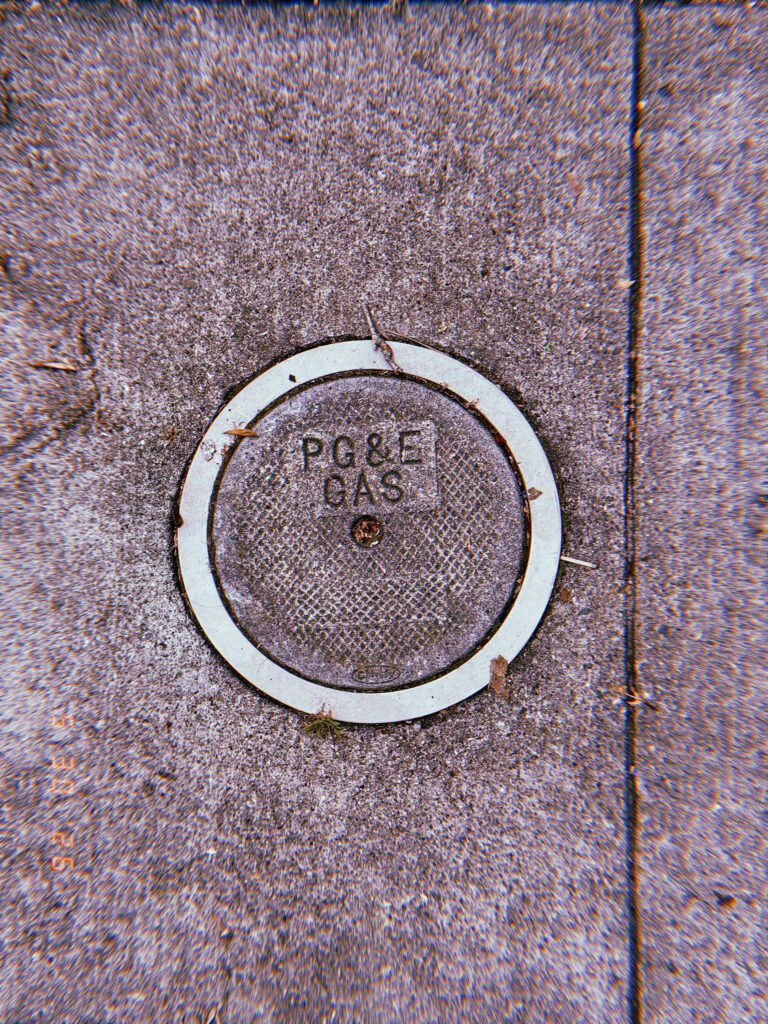 Round PG&E gas utility cover embedded in a textured San Francisco sidewalk, surrounded by a pale circular border and small street debris.