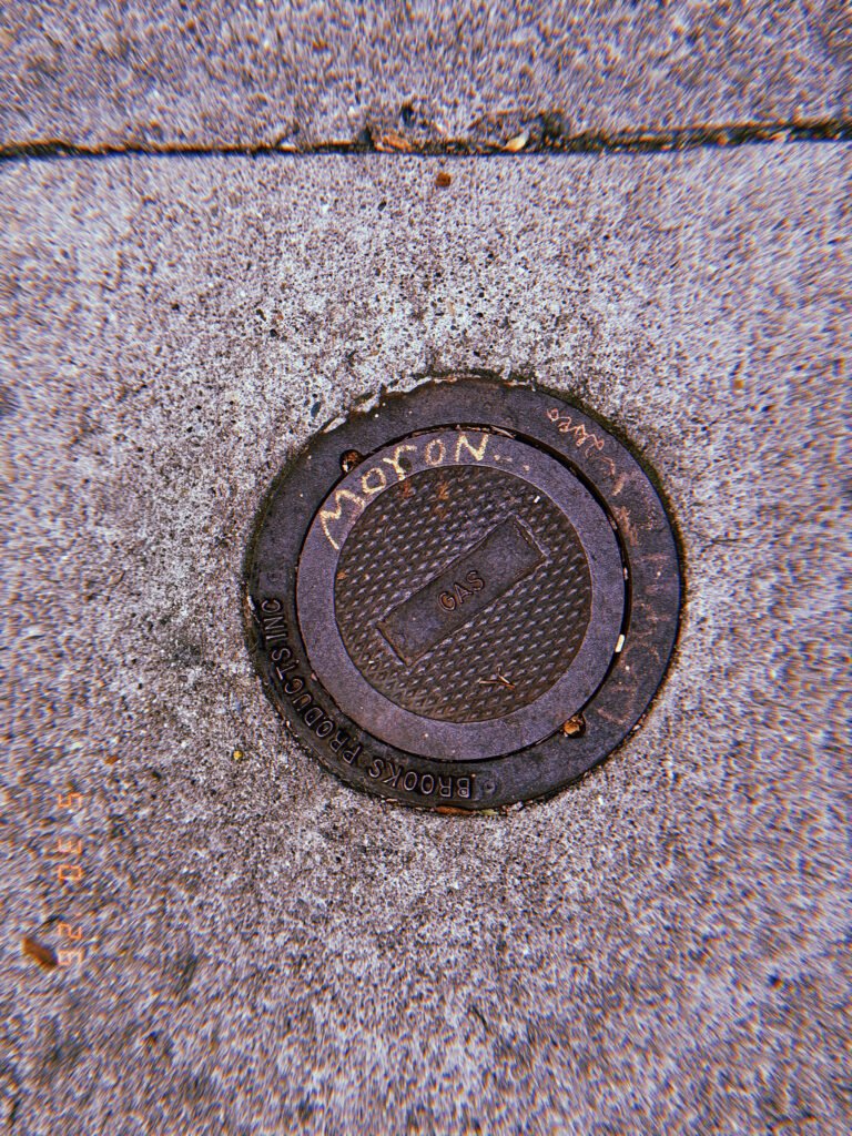 Round metal gas utility valve cover embedded in a concrete San Francisco sidewalk, marked “GAS” and surrounded by worn pavement.