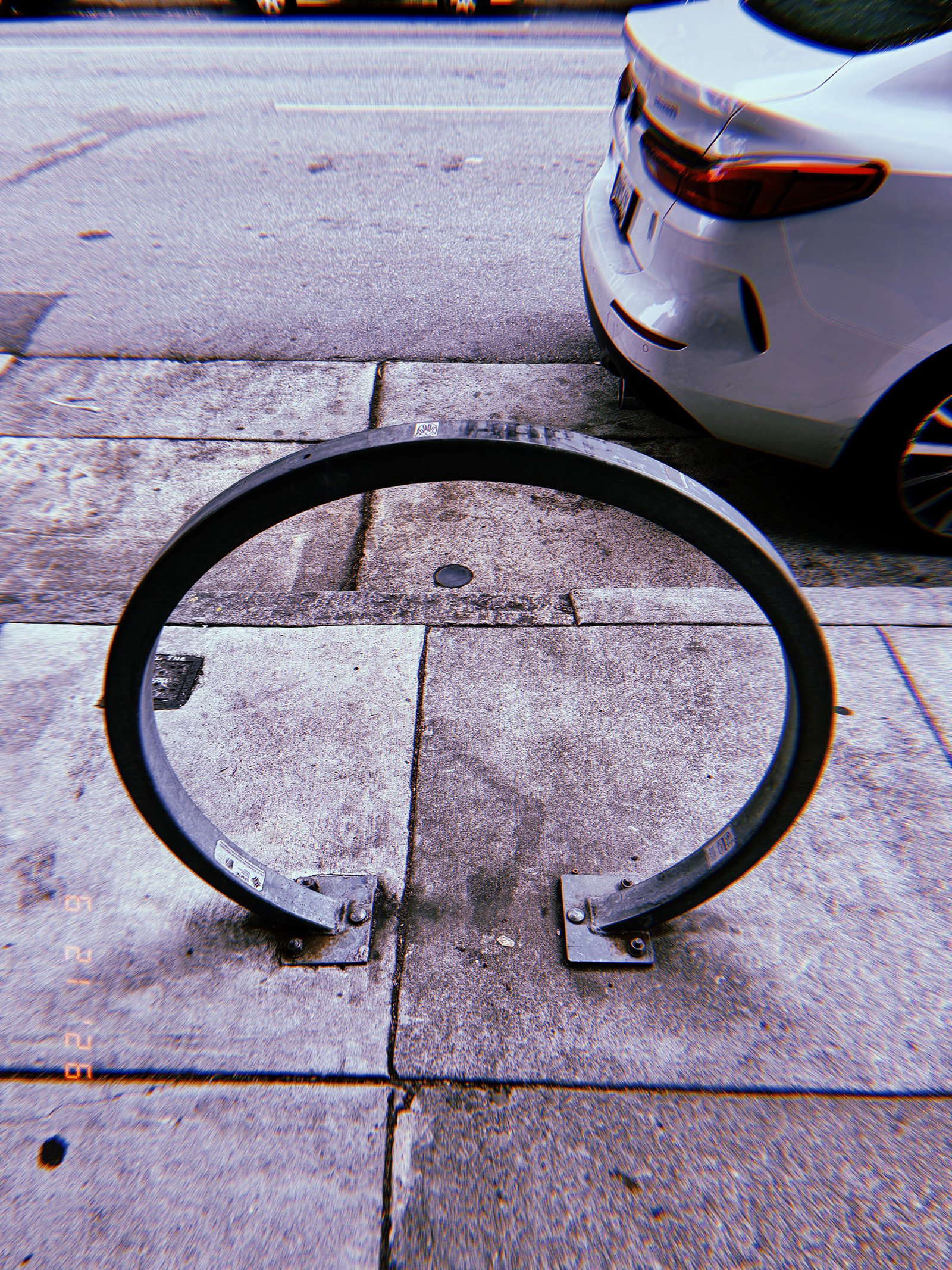 Circular metal bicycle rack mounted on a San Francisco sidewalk beside the curb, with a parked white vehicle and roadway behind it.