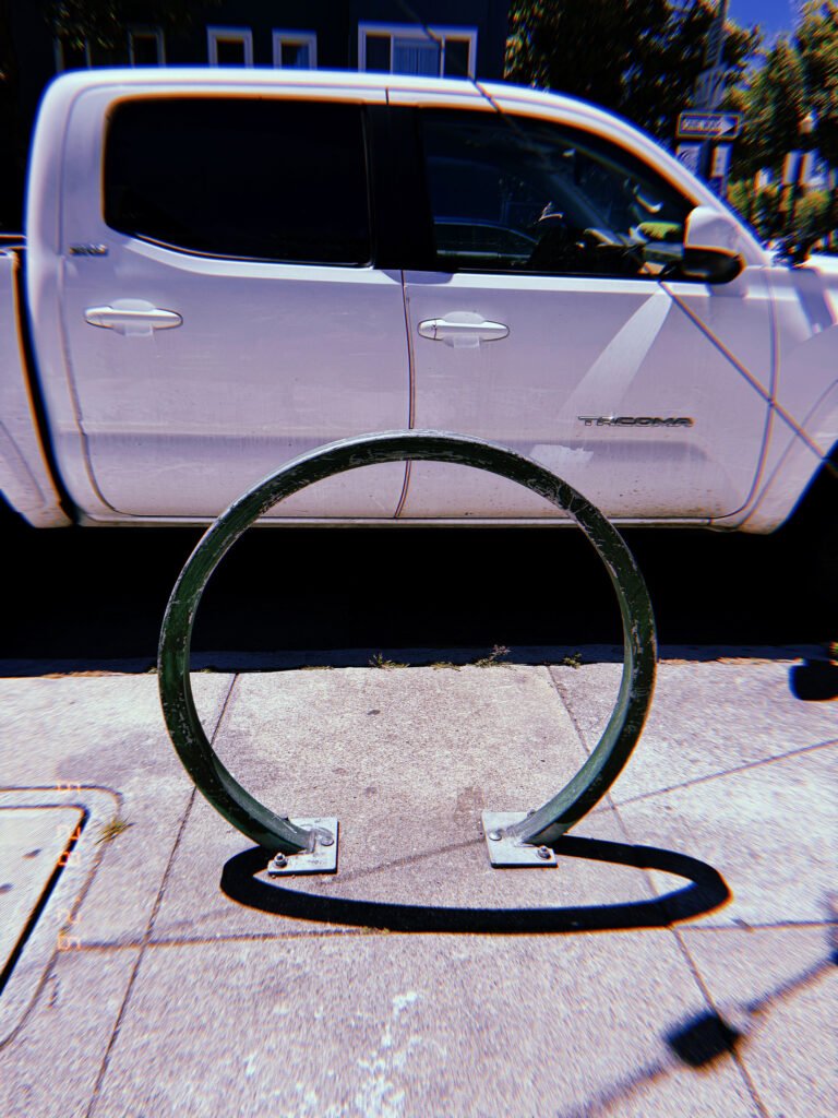 Circular green metal bicycle rack mounted on a San Francisco sidewalk, positioned in front of a parked white pickup truck.