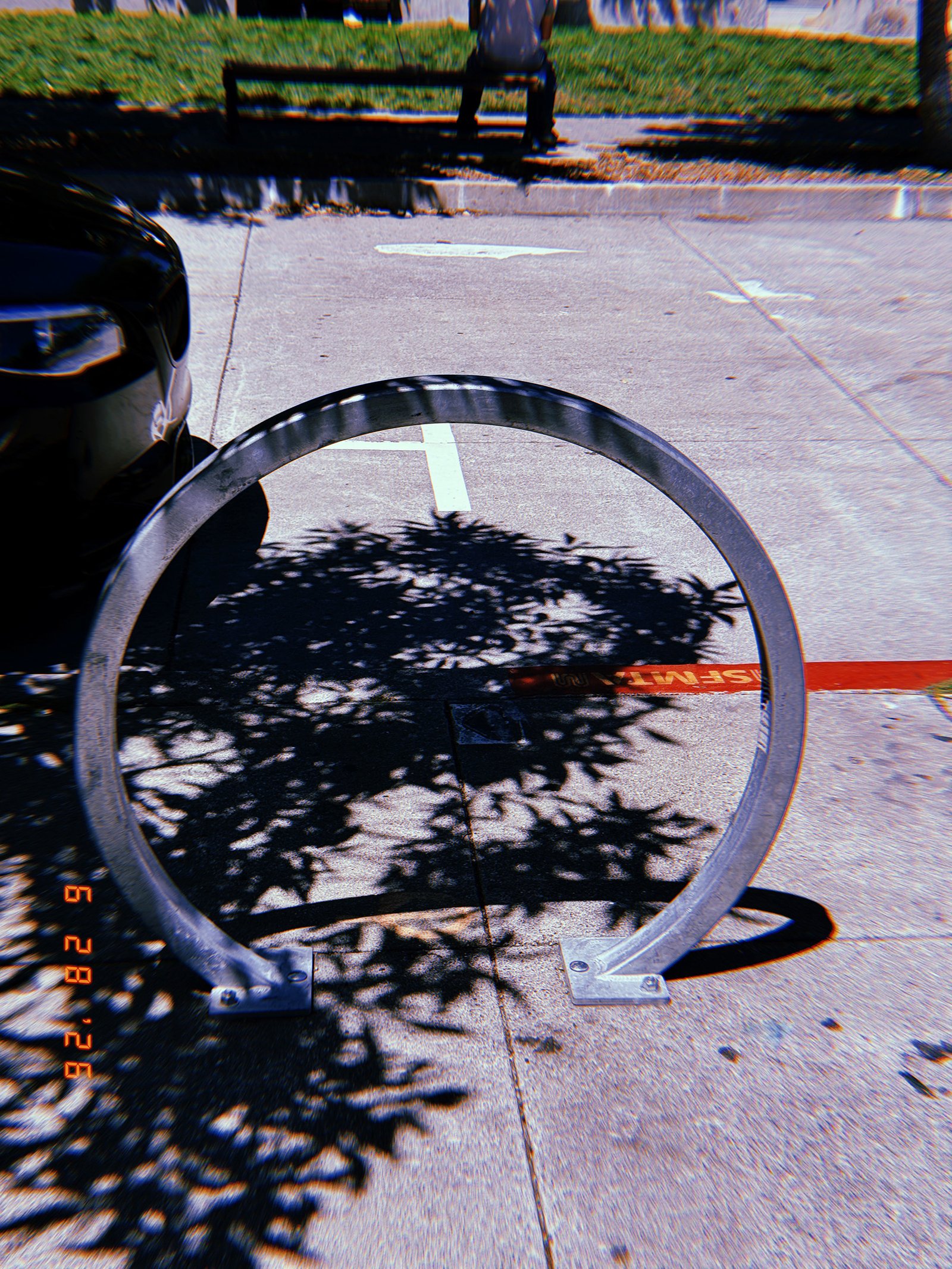 Circular silver metal bicycle rack mounted on a San Francisco sidewalk, with tree shadows, street markings, and a red-painted curb behind it.