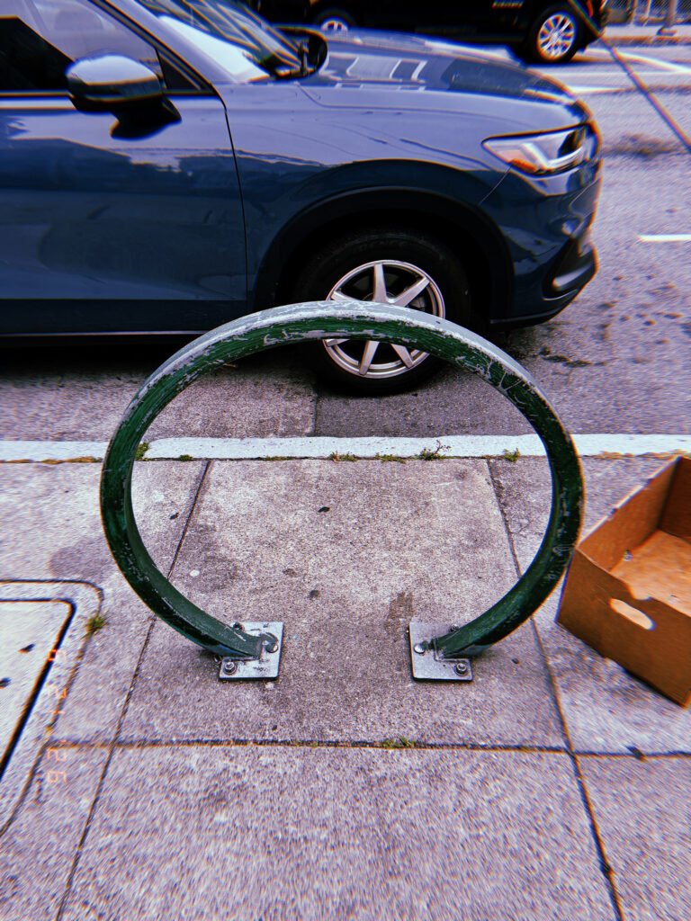 Worn circular green metal bicycle rack mounted on a San Francisco sidewalk, positioned beside a parked blue vehicle.