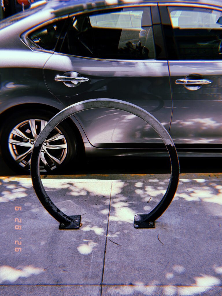 Circular metal bicycle rack mounted on a San Francisco sidewalk, framed against a parked gray SUV with tree shadows across the pavement.