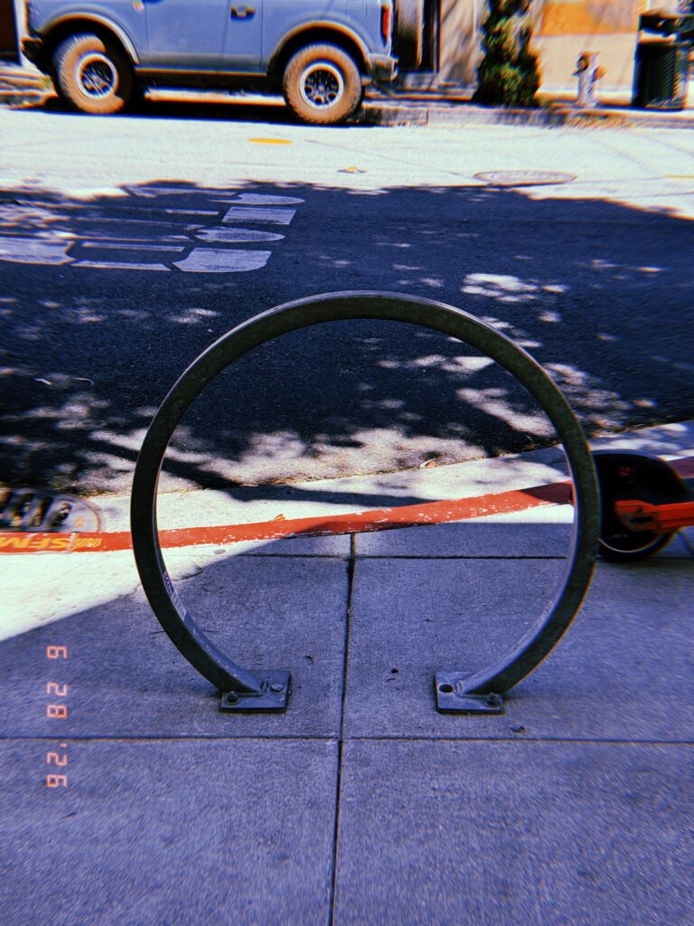 Circular metal bicycle rack mounted on a San Francisco sidewalk beside a red-painted curb, with street markings and parked vehicles behind it.