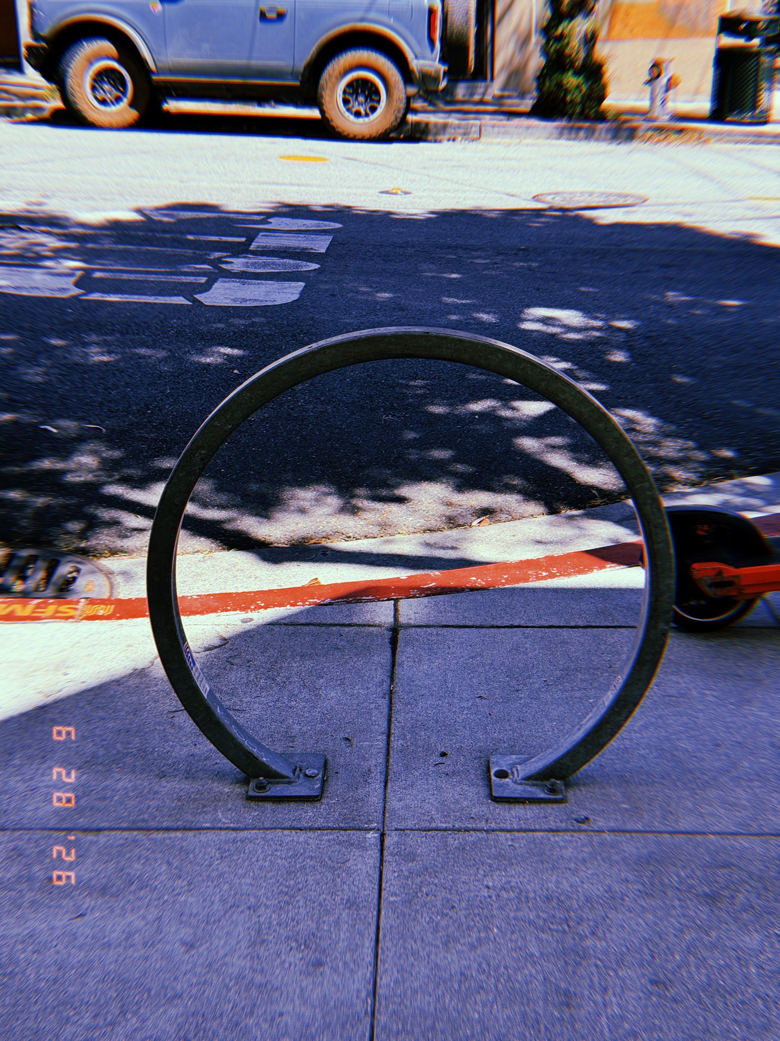 Circular metal bicycle rack mounted on a San Francisco sidewalk beside a red-painted curb, with street markings and parked vehicles behind it.
