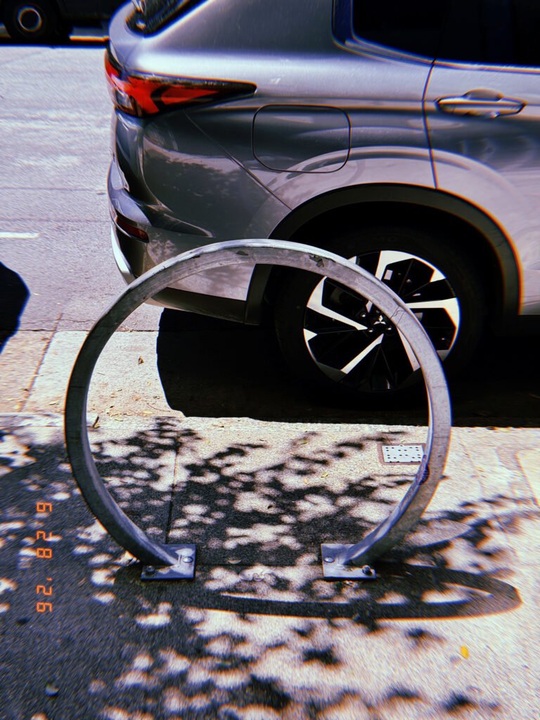 Circular metal bicycle rack mounted on a San Francisco sidewalk, with a parked gray SUV behind it and tree shadows across the pavement.