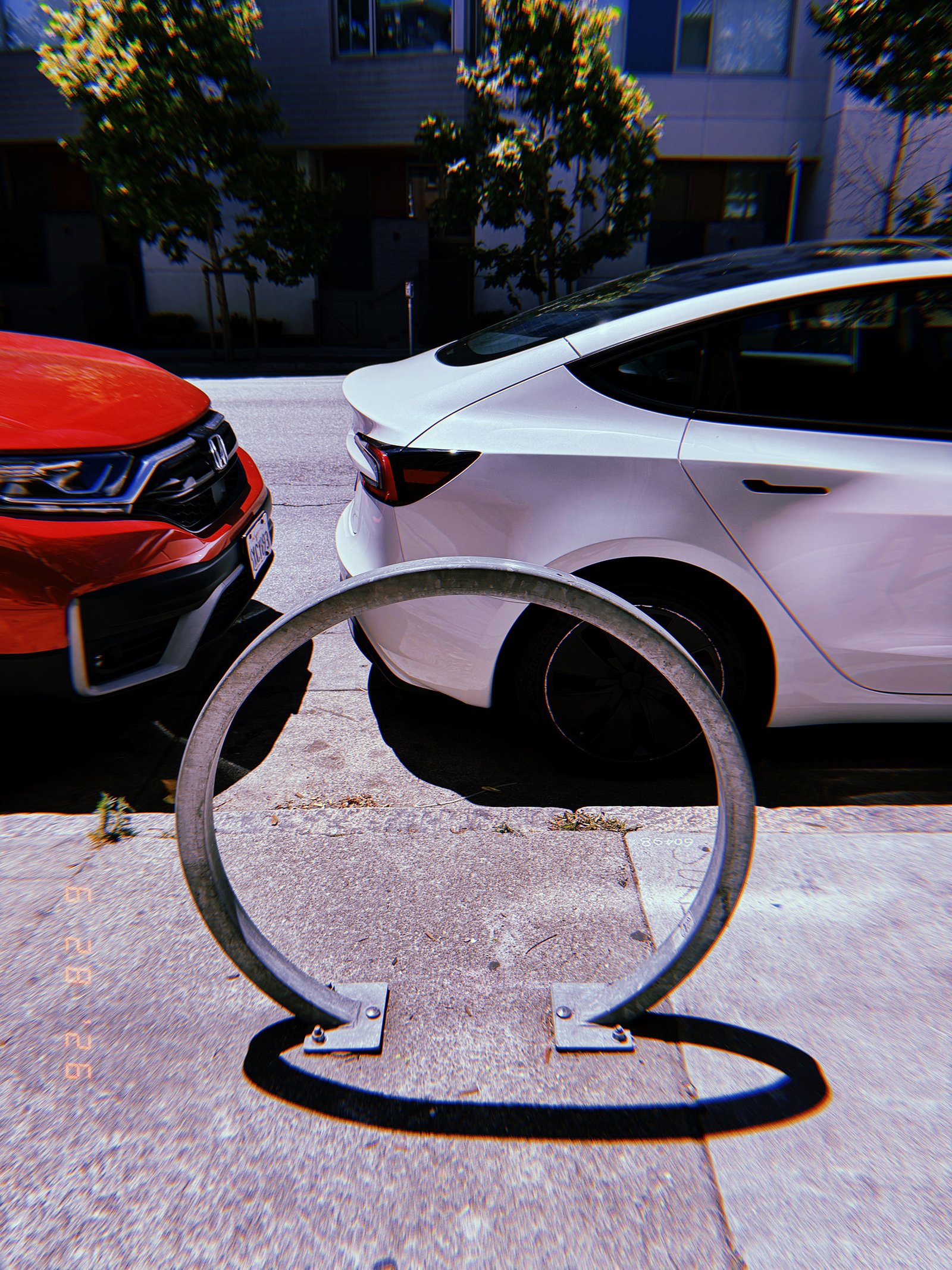 Circular metal bicycle rack mounted on a San Francisco sidewalk, framed against two parked cars and residential buildings.