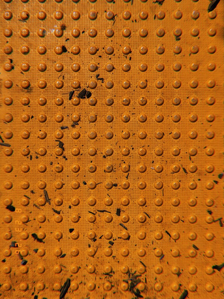 Orange tactile paving with rows of raised truncated domes scattered with dirt, leaves, and small debris.