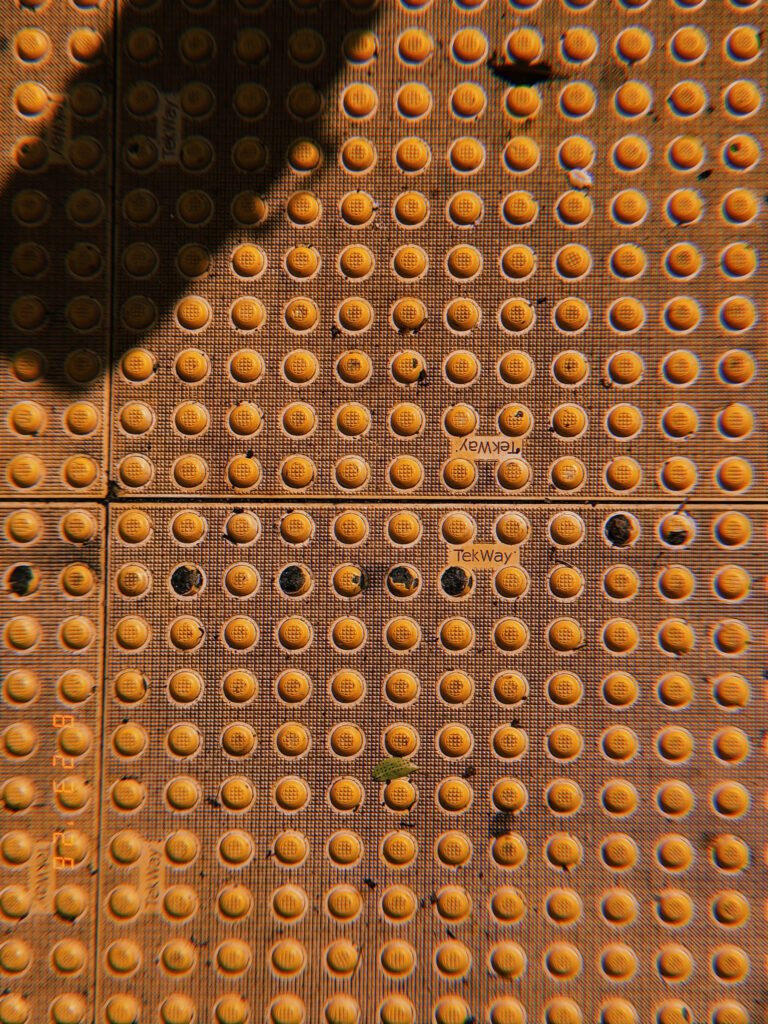 Orange tactile paving with rows of raised truncated domes, worn sections, scattered debris, and strong shadows.