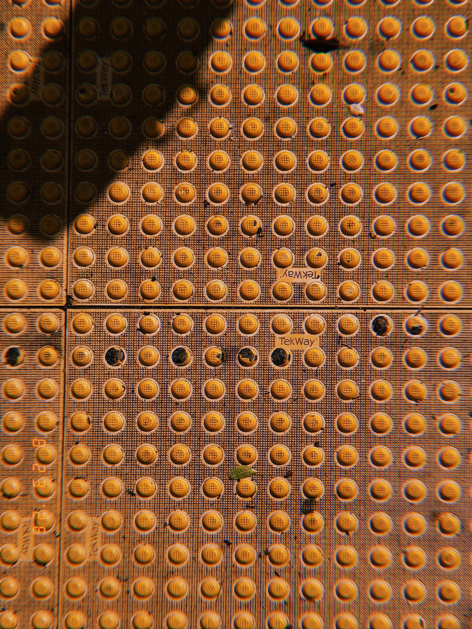 Orange tactile paving with rows of raised truncated domes, worn sections, scattered debris, and strong shadows.
