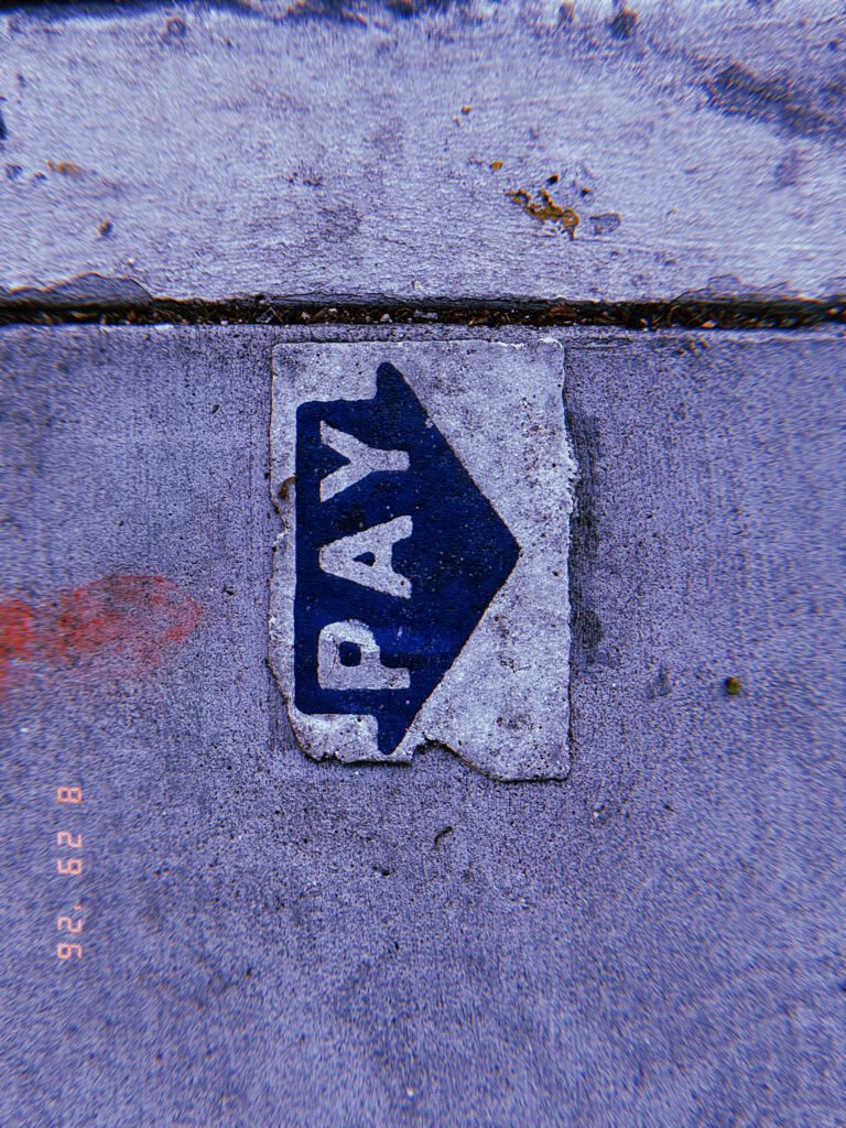 Worn blue PAY arrow marking embedded in a concrete sidewalk, directing pedestrians toward a parking payment point.