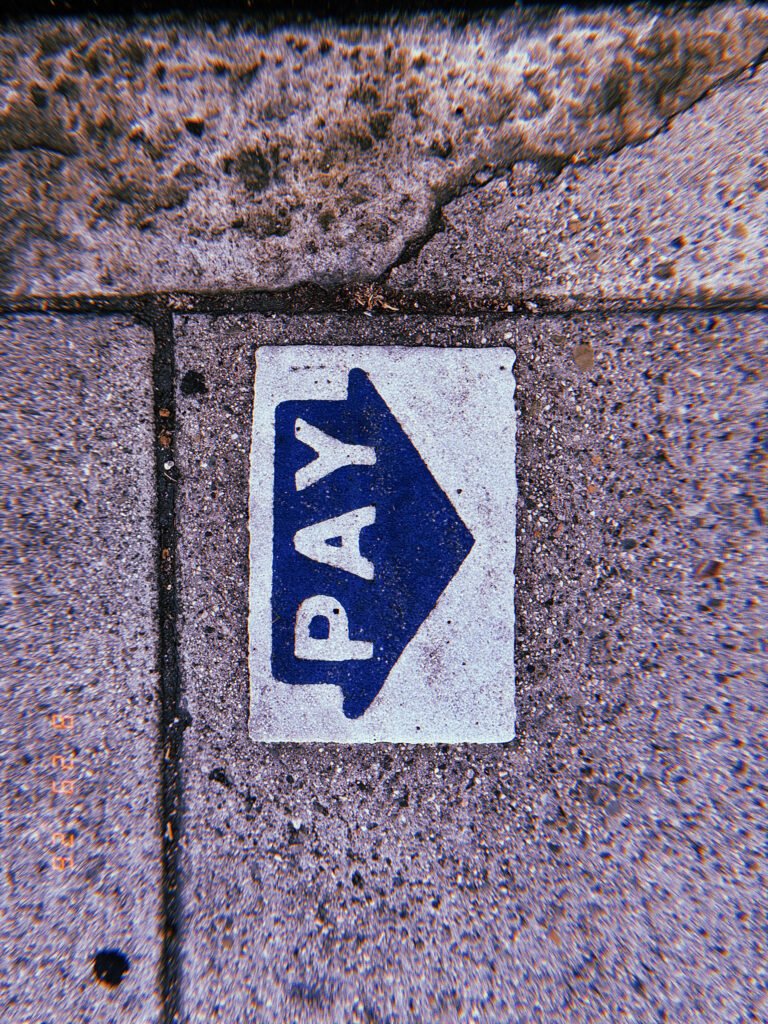 Blue PAY arrow marking embedded in a worn concrete sidewalk, directing pedestrians toward a parking payment point.