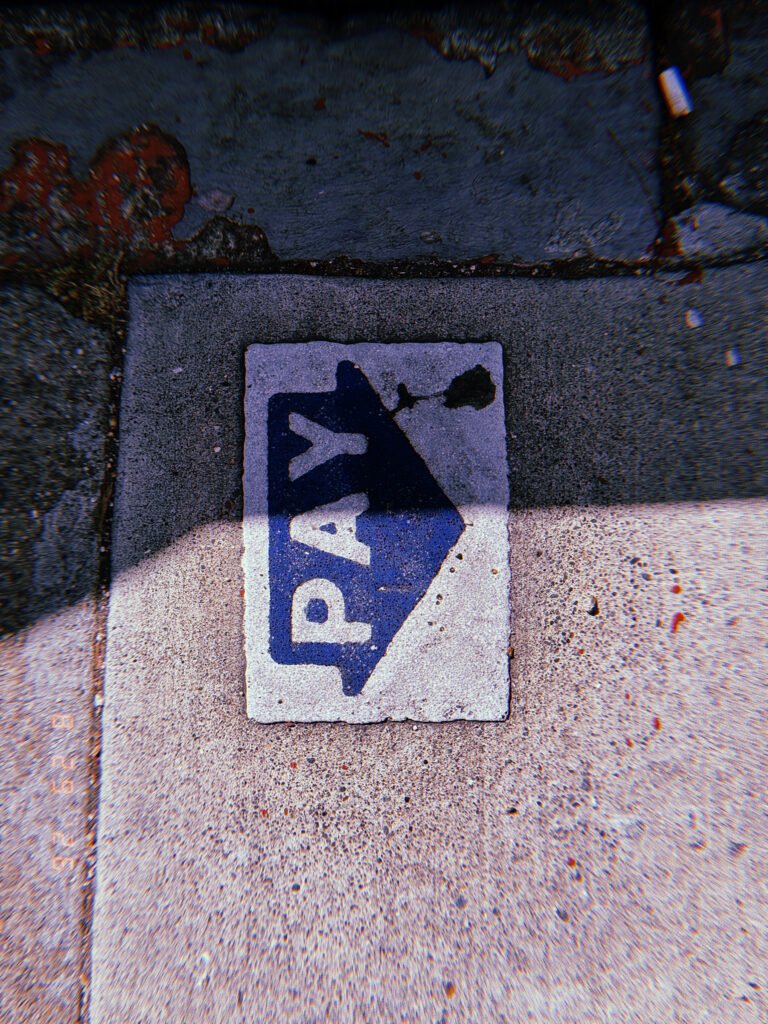 Blue PAY arrow embedded in a concrete sidewalk, partially crossed by a strong shadow beside a worn painted curb.