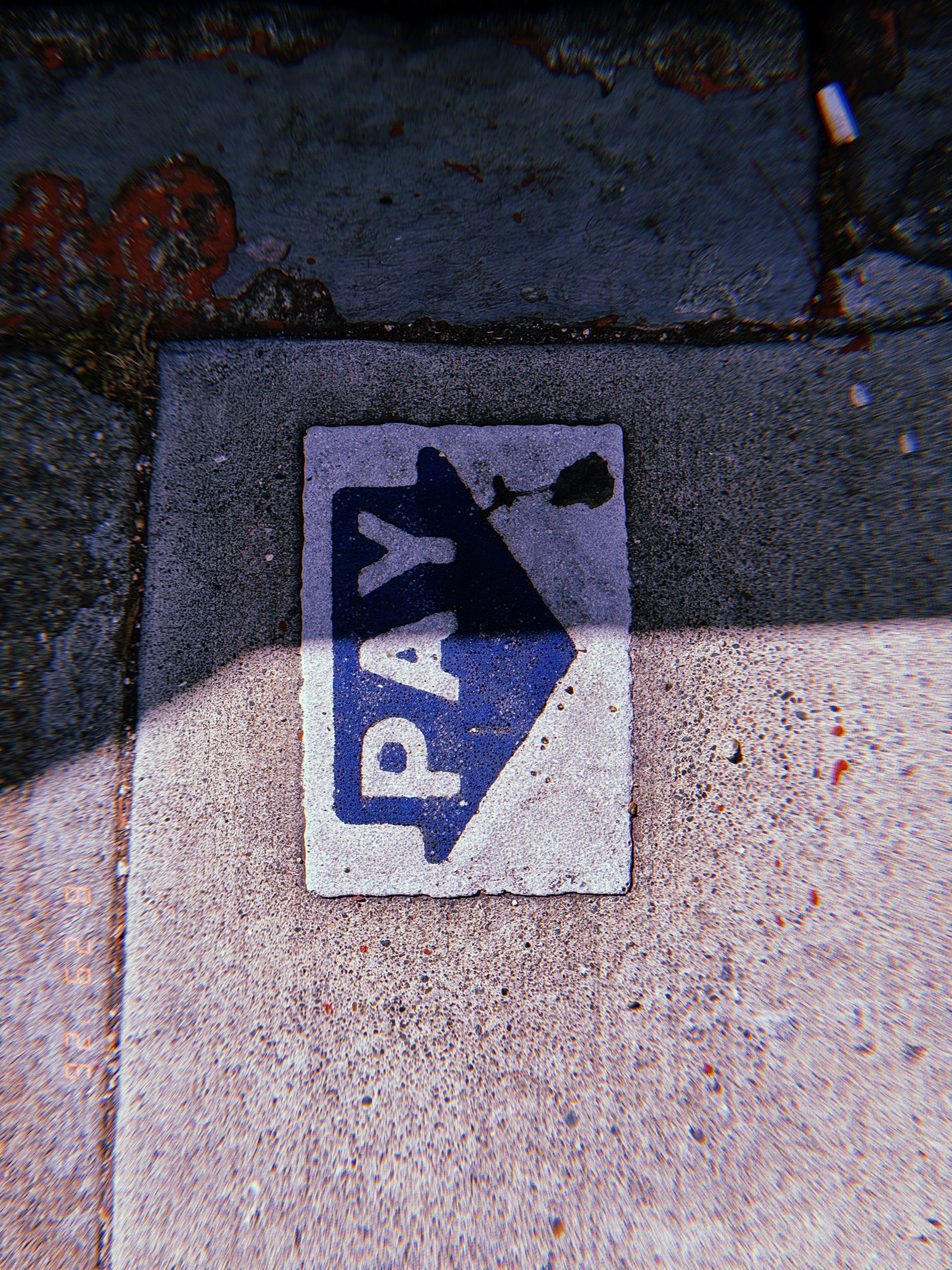 Blue PAY arrow embedded in a concrete sidewalk, partially crossed by a strong shadow beside a worn painted curb.