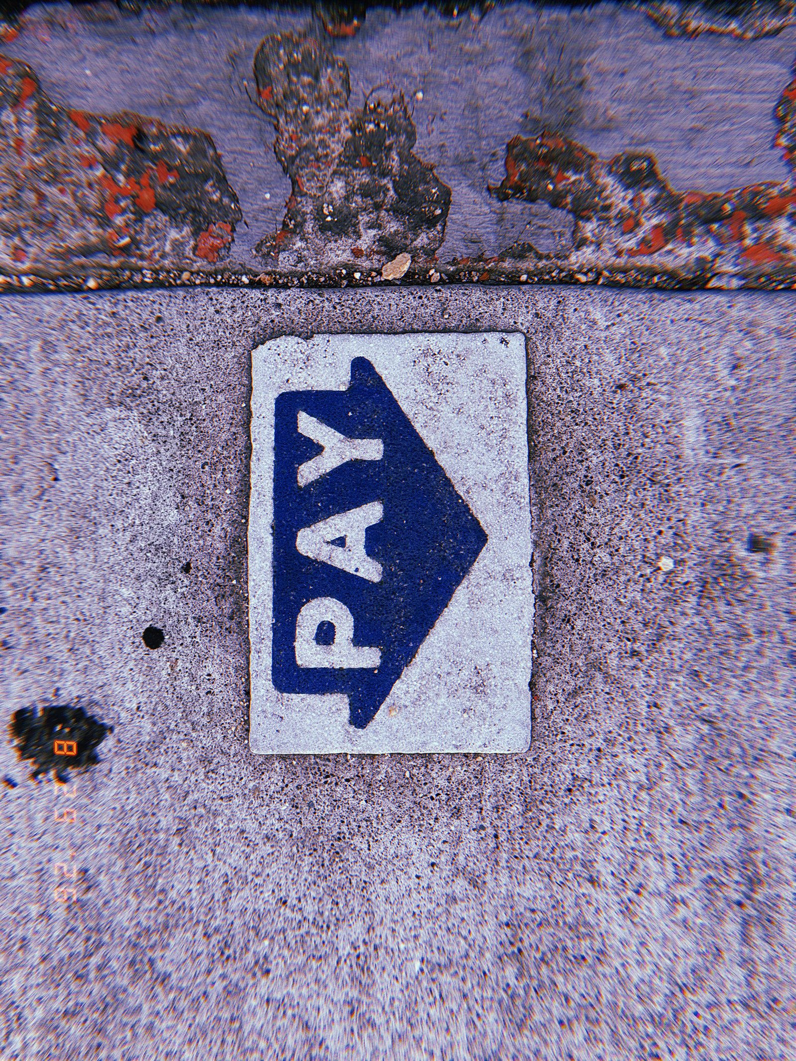 Blue PAY arrow marking embedded in a concrete sidewalk beside a worn painted curb.