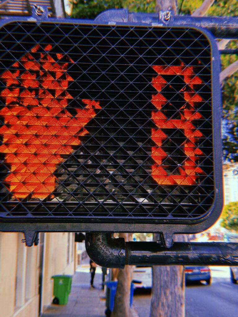 Red pedestrian stop-hand symbol and countdown number 9 illuminated behind a black geometric protective grid on a crossing signal in San Francisco.
