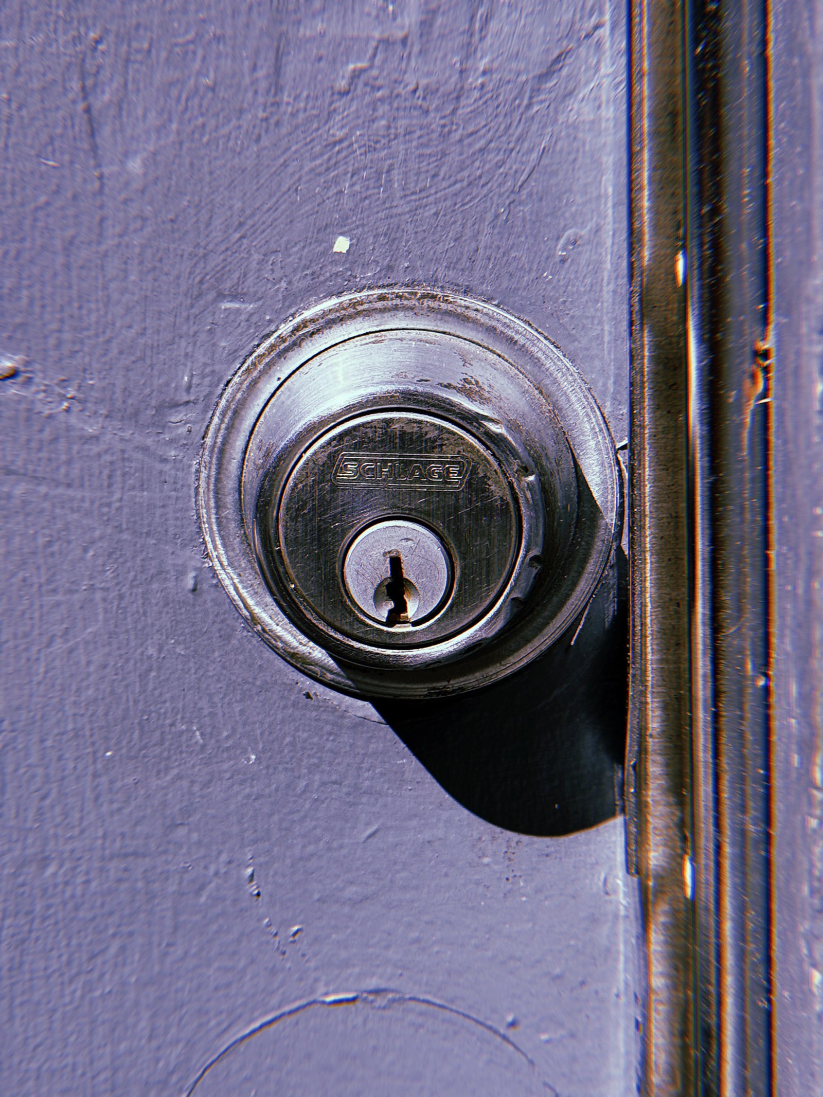 Dark metal Schlage deadbolt lock mounted on a muted purple painted door, with scratched metal surfaces, worn edges, and a curved shadow beneath the lock.